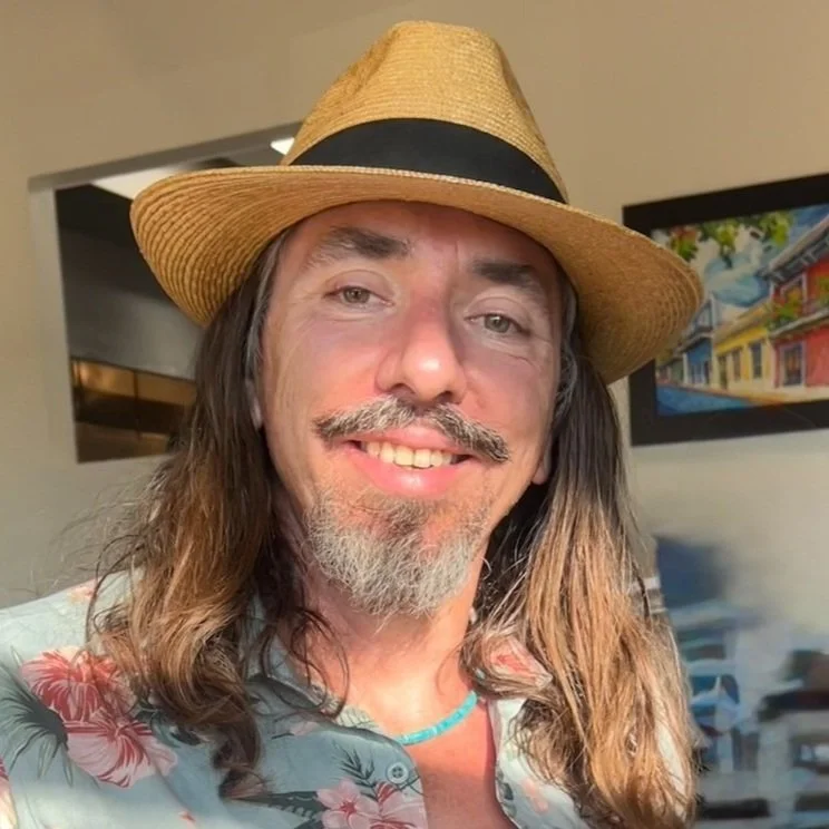 Man with long brown hair, a gray beard, and mustache, wearing a straw hat with a black band, a floral shirt, and a turquoise necklace, smiling indoors in front of a colorful painting.
