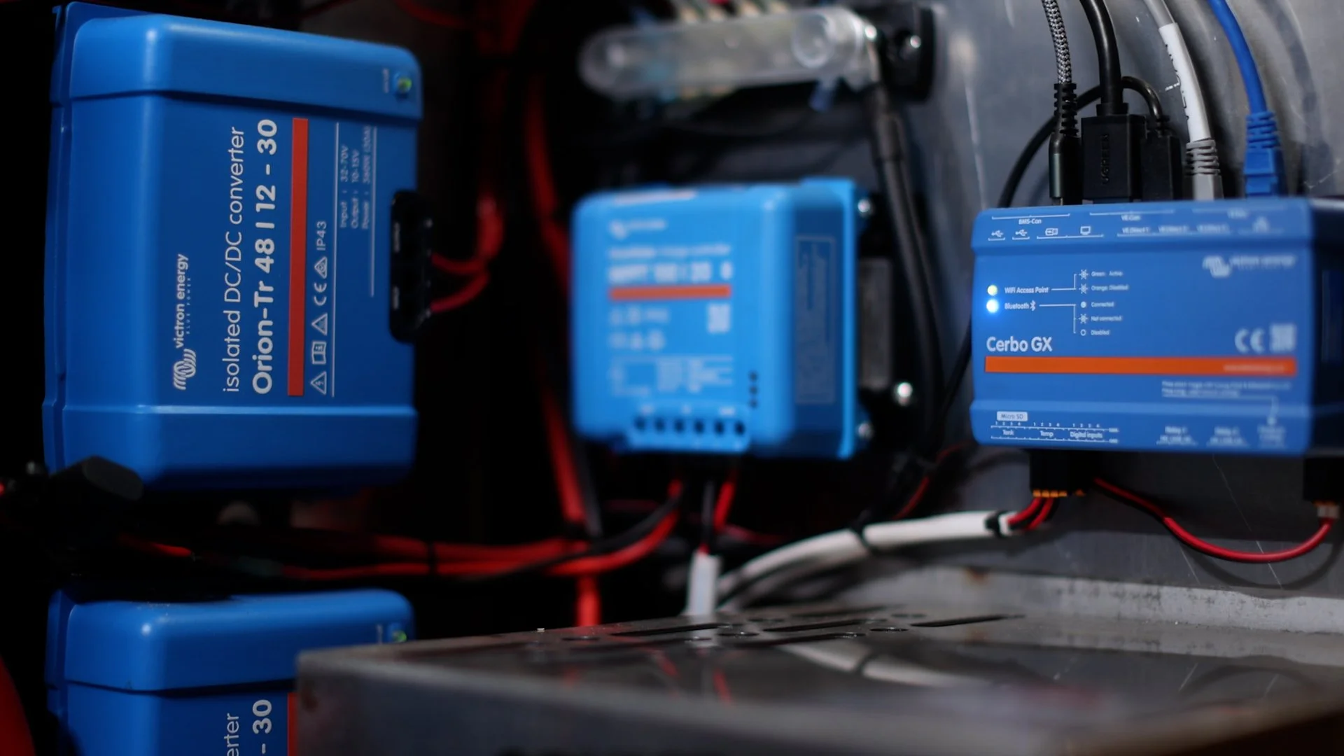 Close-up of electrical components, including blue Victron Orion-TR 48/12-30 inverter, a blue Cerbo GX device, and other electrical modules with wires connected, mounted on a metal surface.