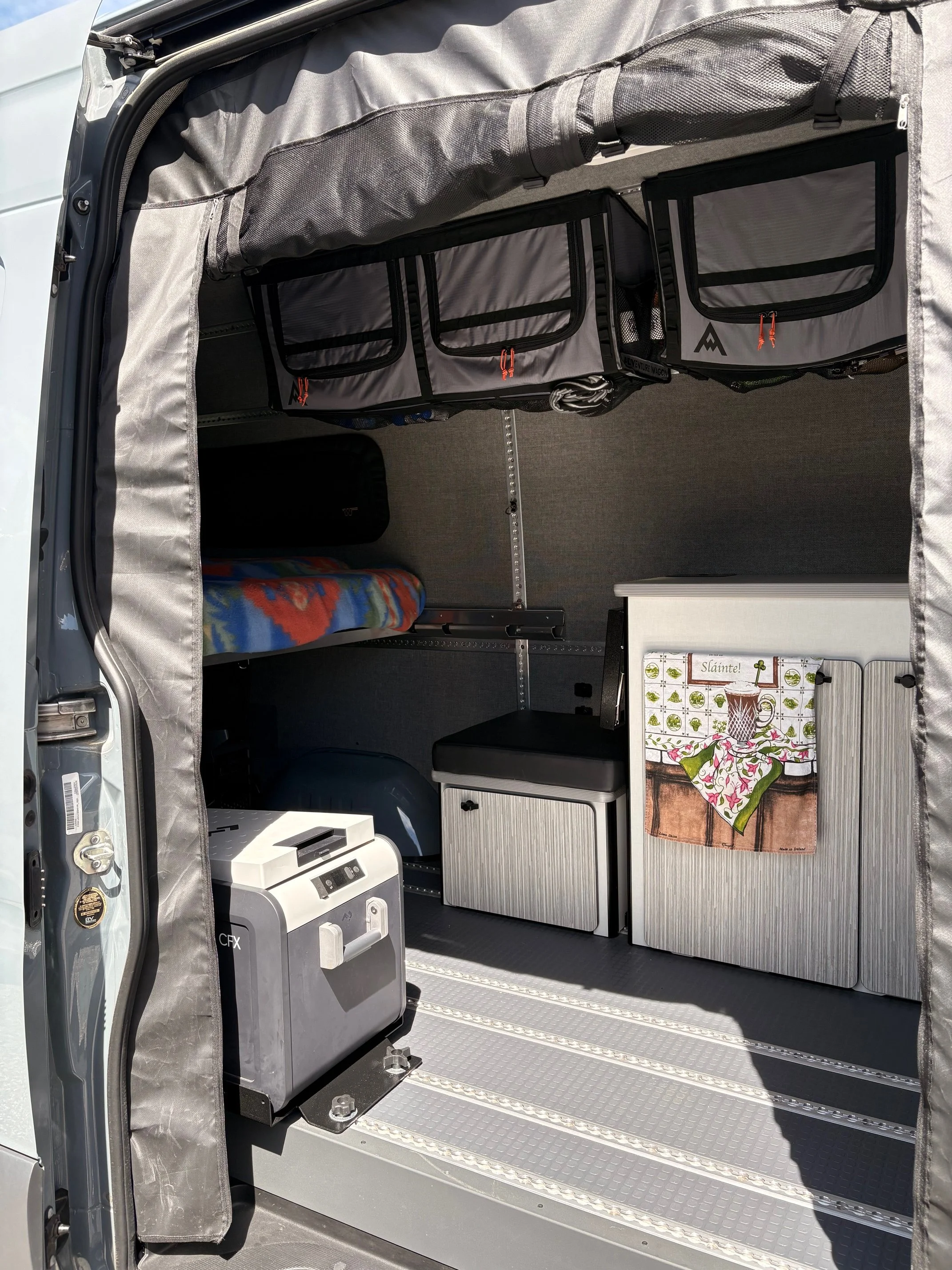 The interior of a van converted into a camper with storage compartments, a small table with a decorative cloth, and various supplies including a portable generator and a blanket.