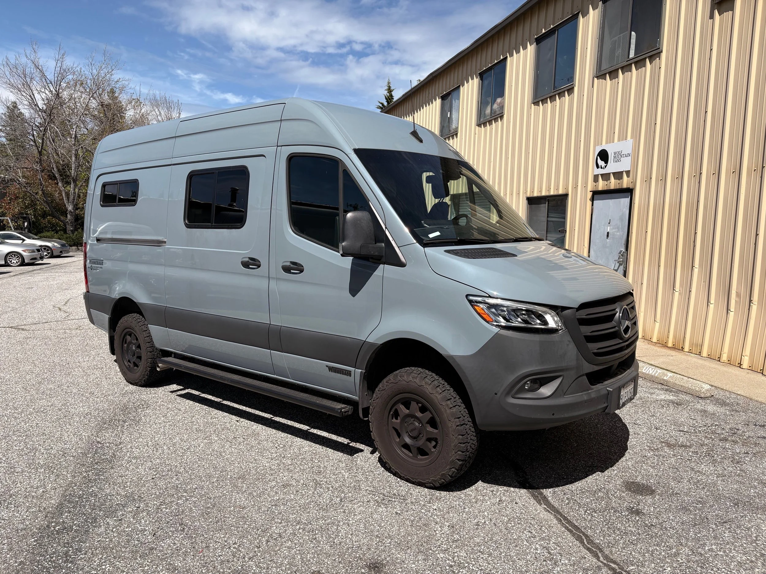 A silver Mercedes-Benz Sprinter van parked in front of a beige metal building with a sign that reads 'Wolf Mountain Vans'.