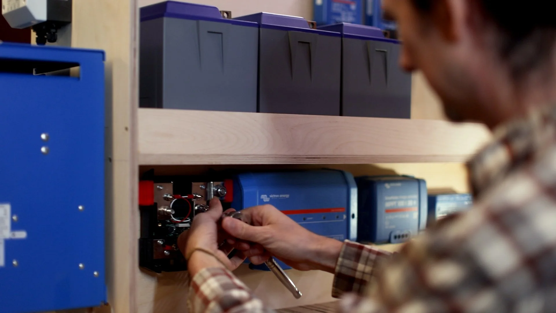 Person working on electrical equipment on a wooden shelf, with blue and black circuit boxes behind.