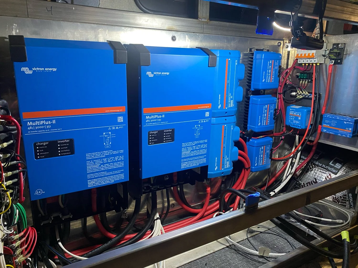 Electrical components and wiring inside a metallic enclosure, including blue Victron energy MultiPlus-II inverters, smaller blue batteries, and miscellaneous electrical parts.