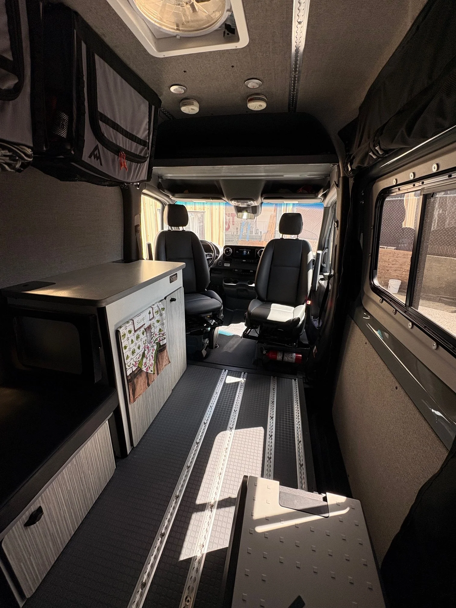 Inside the front cabin of a camper van with two driver seats, a small kitchenette with a countertop and cabinets, and storage compartments visible above and around the seats. The van has windows on the side and front, and the ceiling features smoke detectors and a mounted vent.