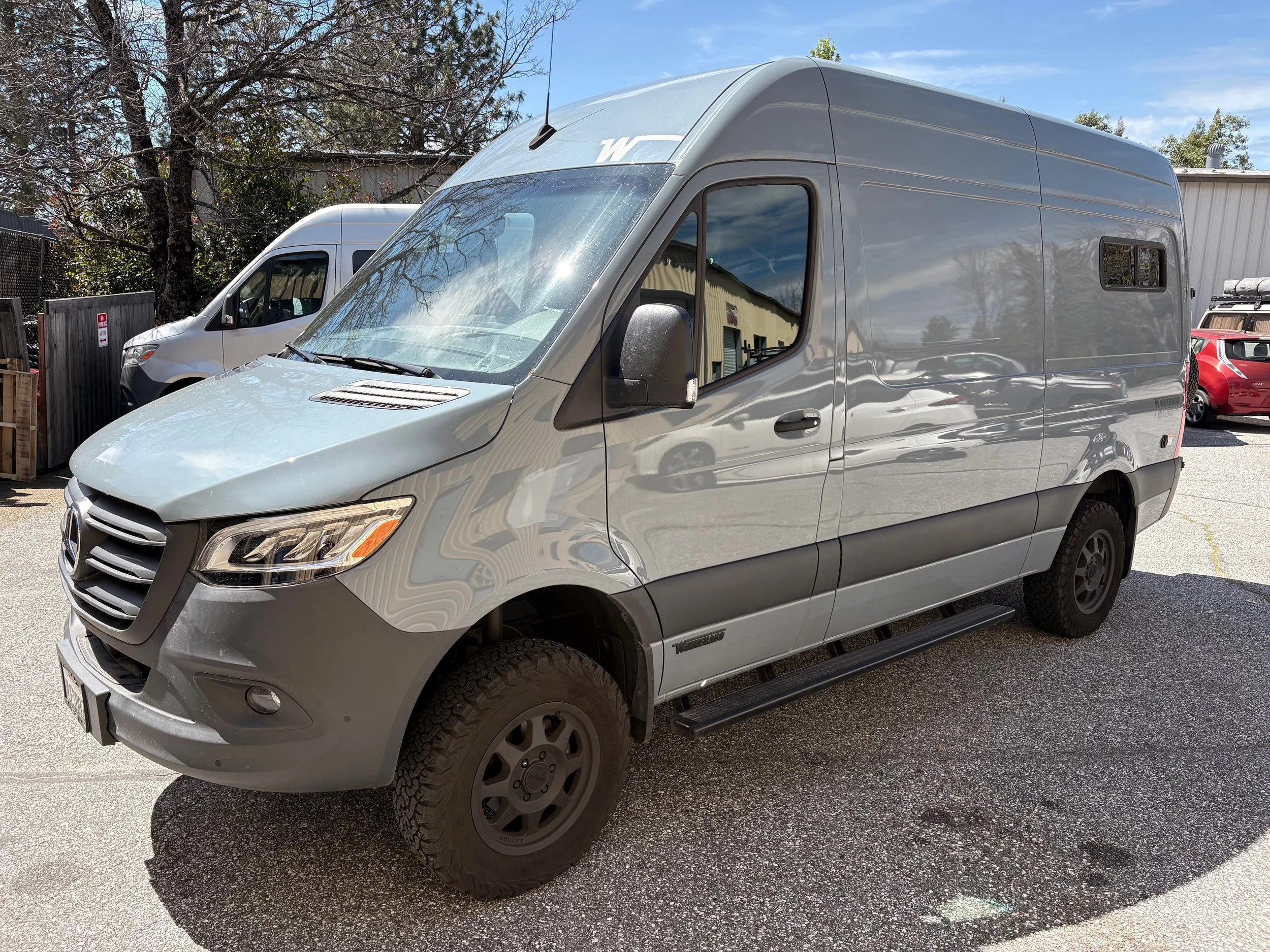 Gray camper van parked outdoors with other vehicles and trees in the background on a sunny day.