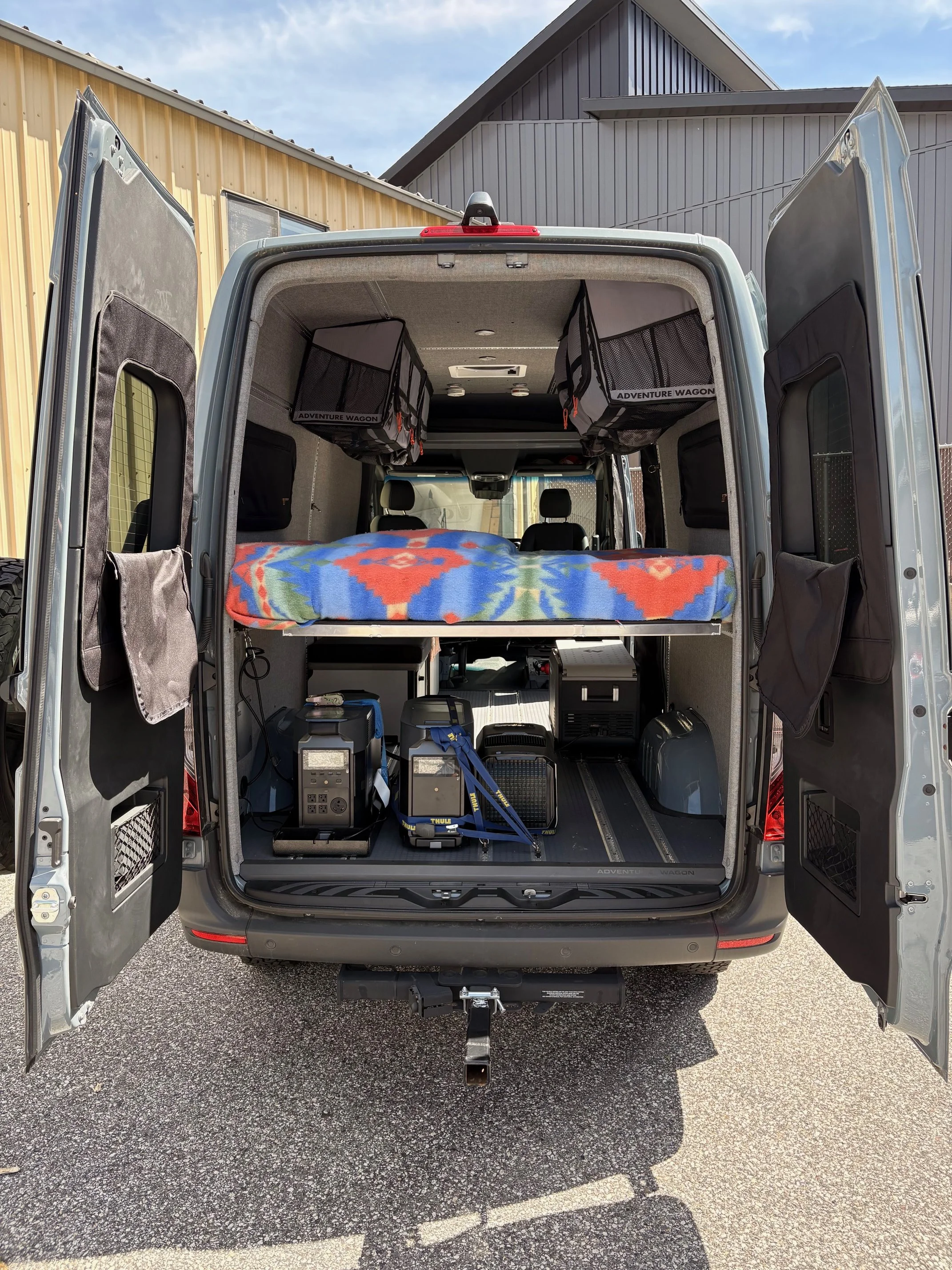 Open van with interior showing storage compartments and a bed with a blanket, set in a driveway outside houses.