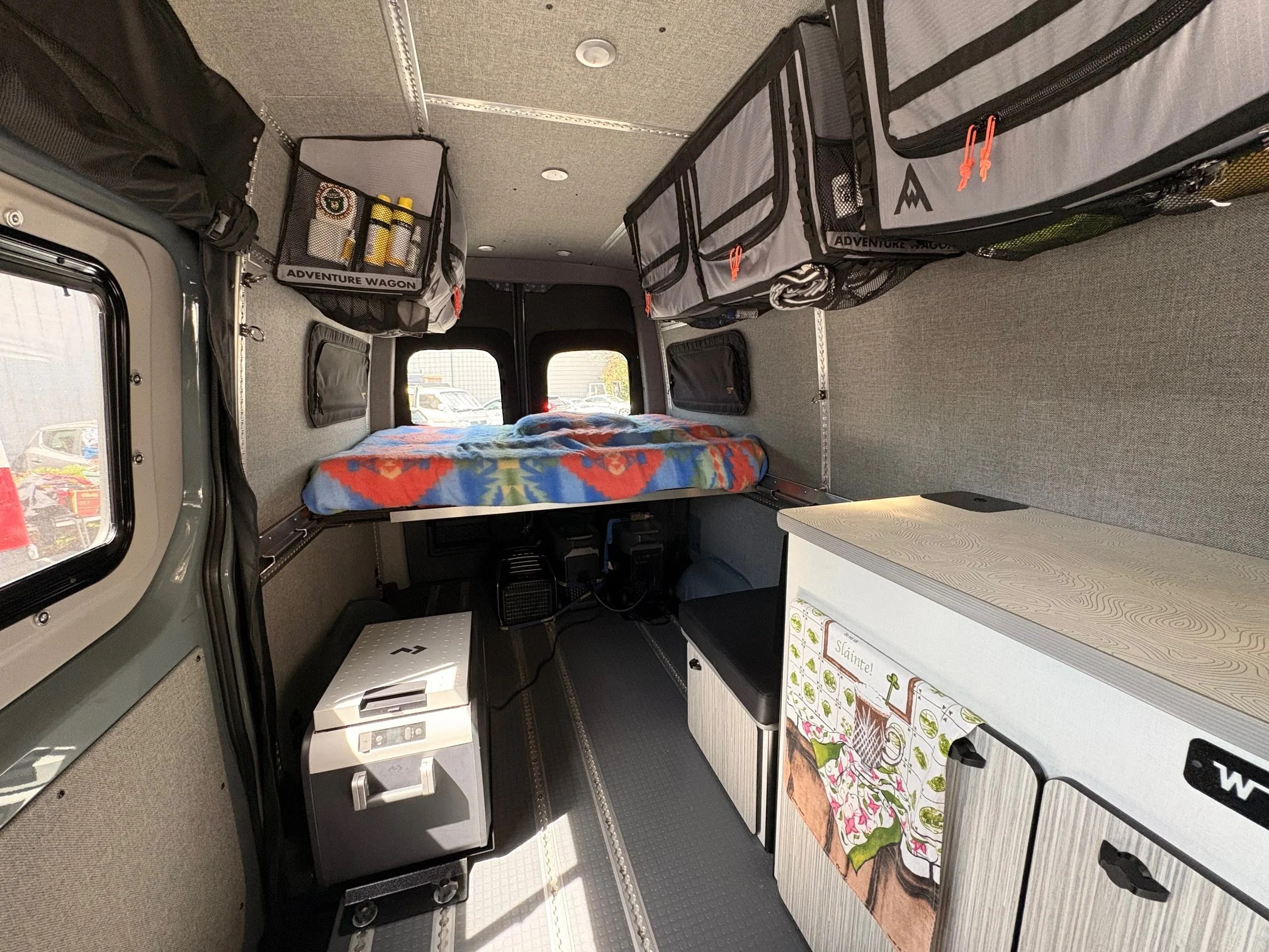 Interior of a camper van or small RV, showing a bed with a colorful blanket, storage compartments labeled 'Adventure Wagon,' a small table with a floral tablecloth, and various appliances and storage areas.