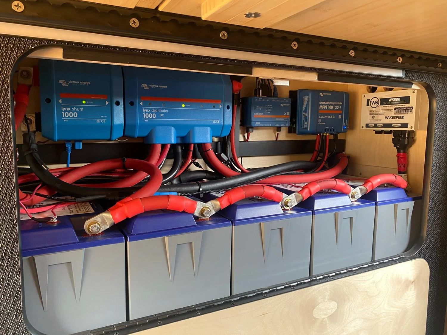 An electrical battery system with four large batteries, red and black cables, and blue controllers inside a wooden enclosure.
