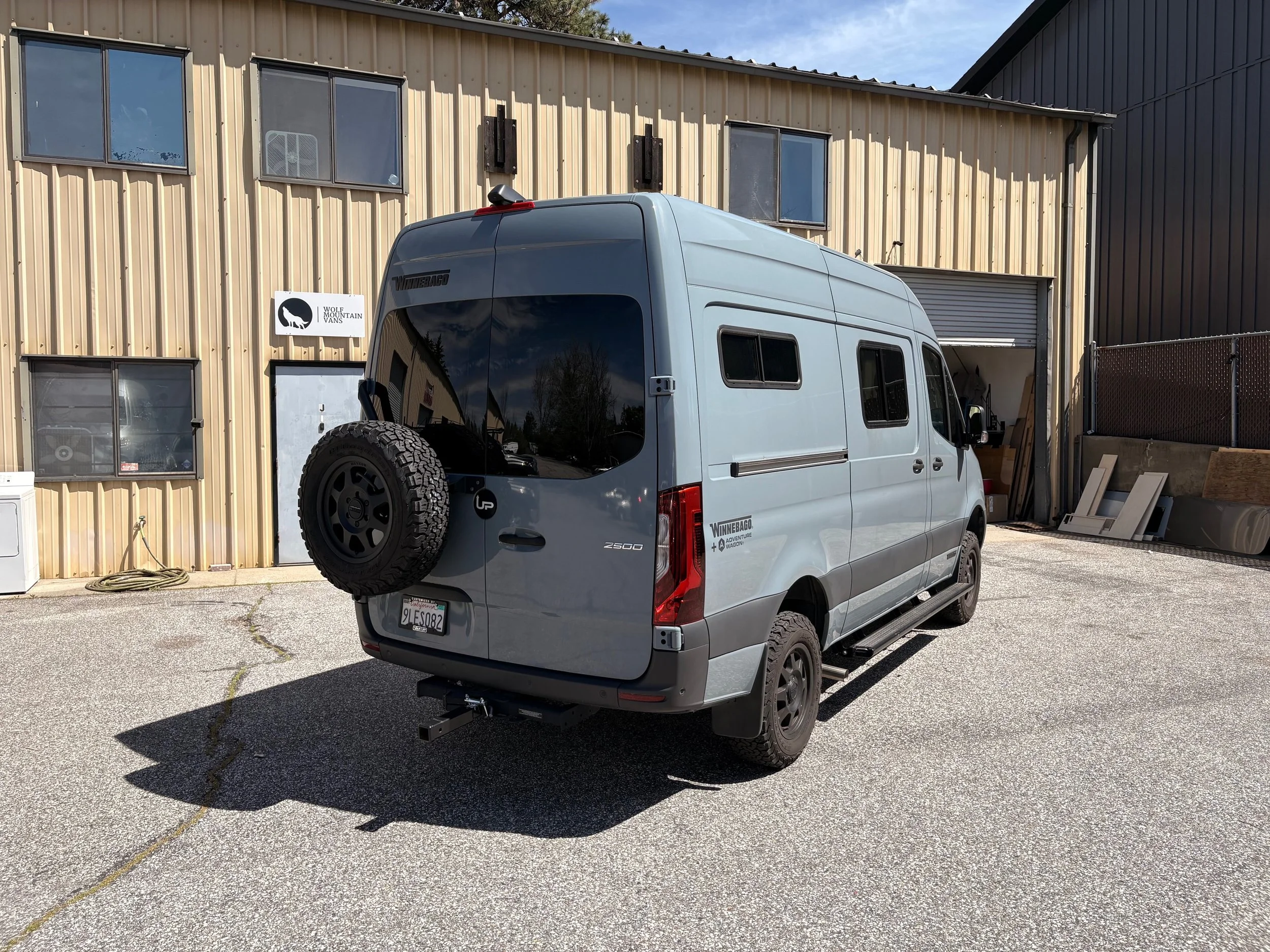 A white Winnebago adventure wagon camper van parked in front of a beige metal building with several windows and a Wolf Mountain Vans sign. The sky is partly cloudy and there are trees reflected in the van's windows.