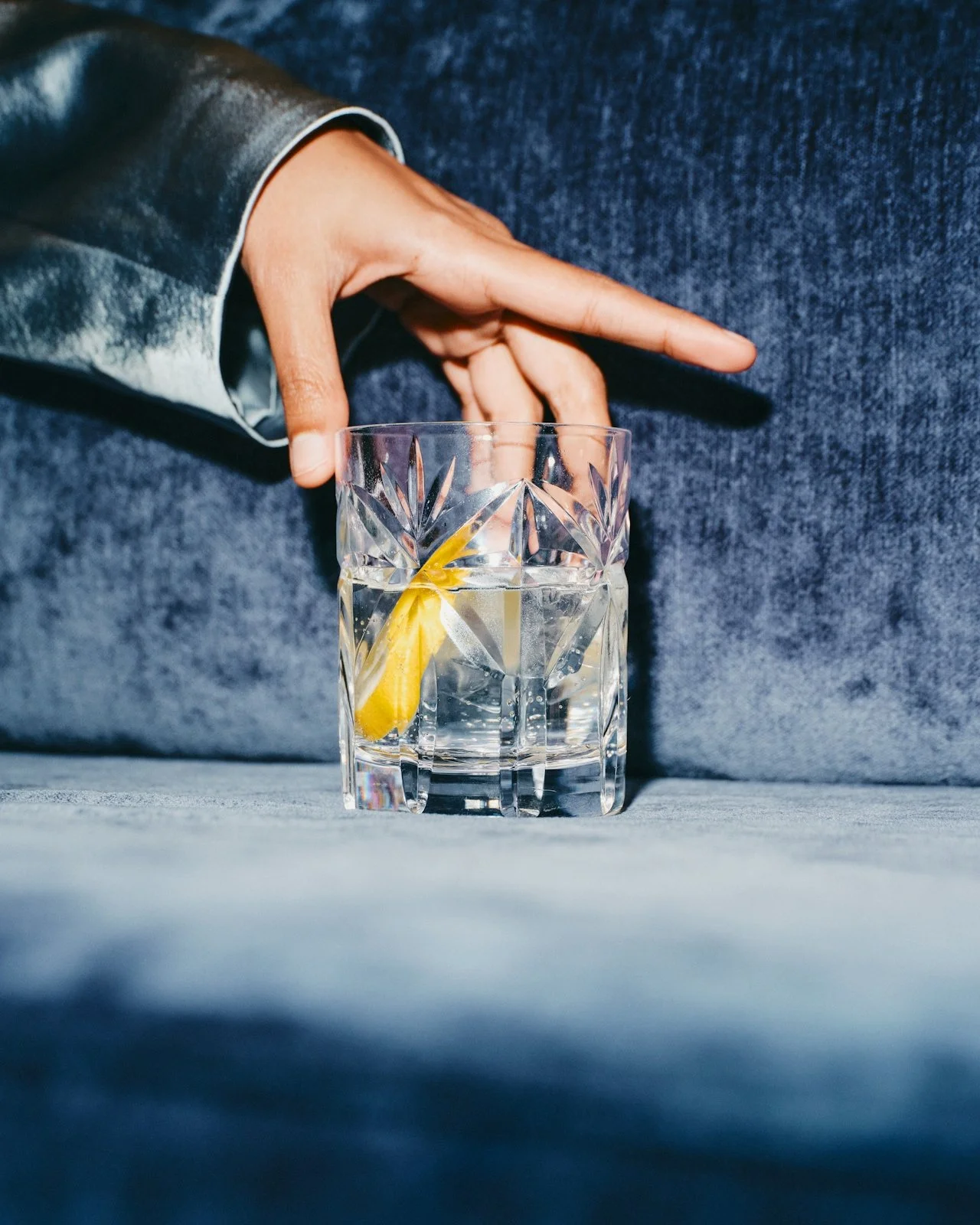 A hand reaching for a glass of water with a lemon wedge on a blue fabric surface.