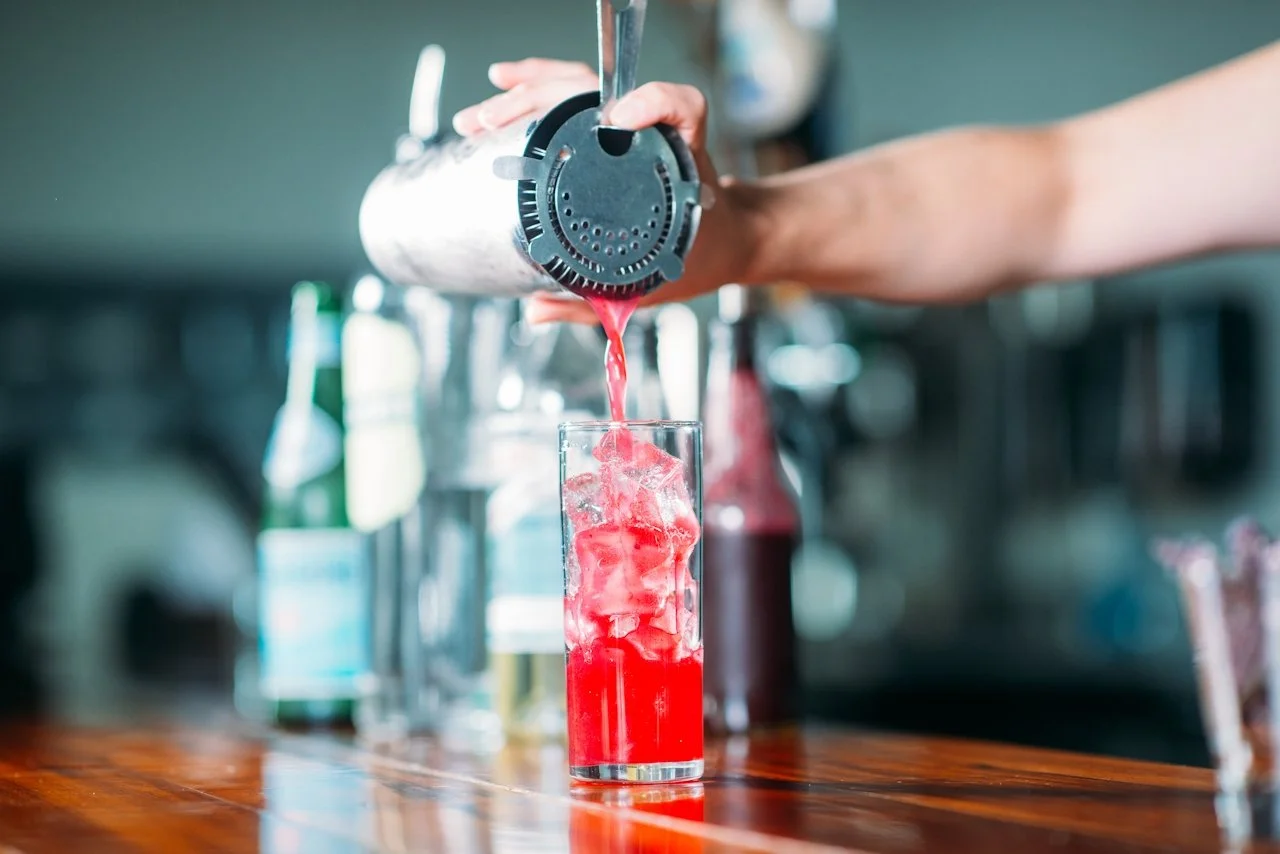 A person pouring a red beverage over ice into a tall glass, with a soda gun on a bar or kitchen counter.