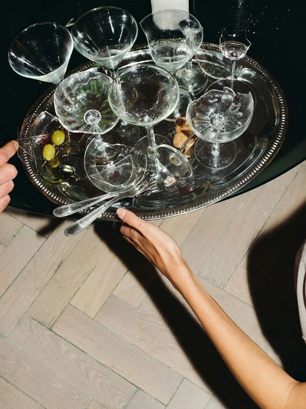 A metal tray with empty glassware including wine glasses and cocktail glasses, two green grapes, cork, a corkscrew, and metal utensils, being carried by two hands.