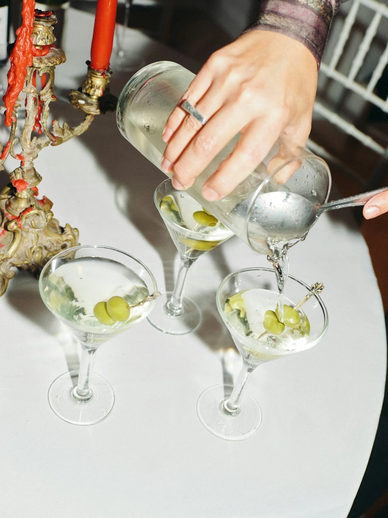 A person pours a clear beverage with green olives into three cocktails, each garnished with green olives, on a white table.