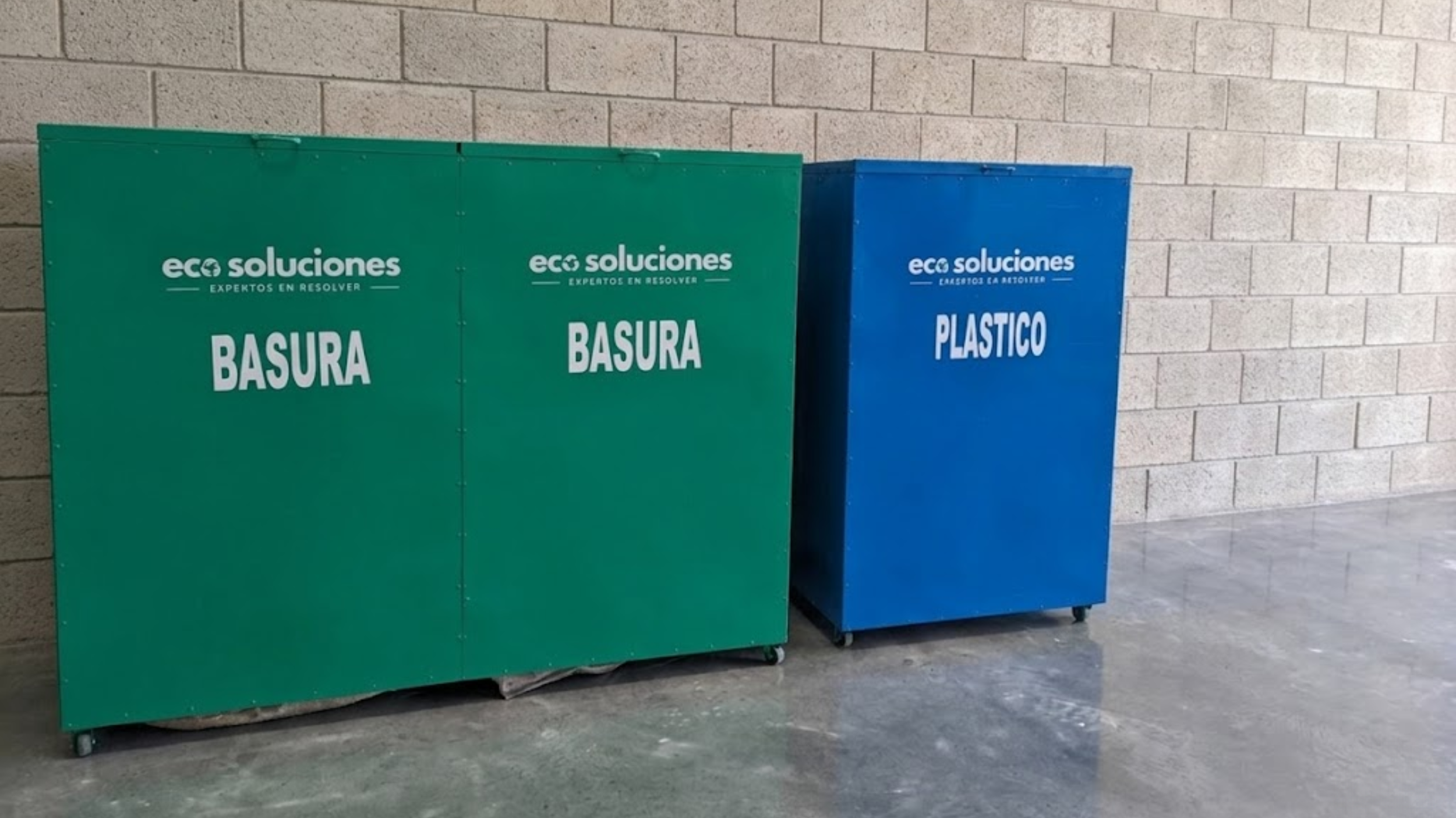 Contenedores de basura verde y azul con etiquetas en español, ubicados en un pasillo con pared de bloques de concreto