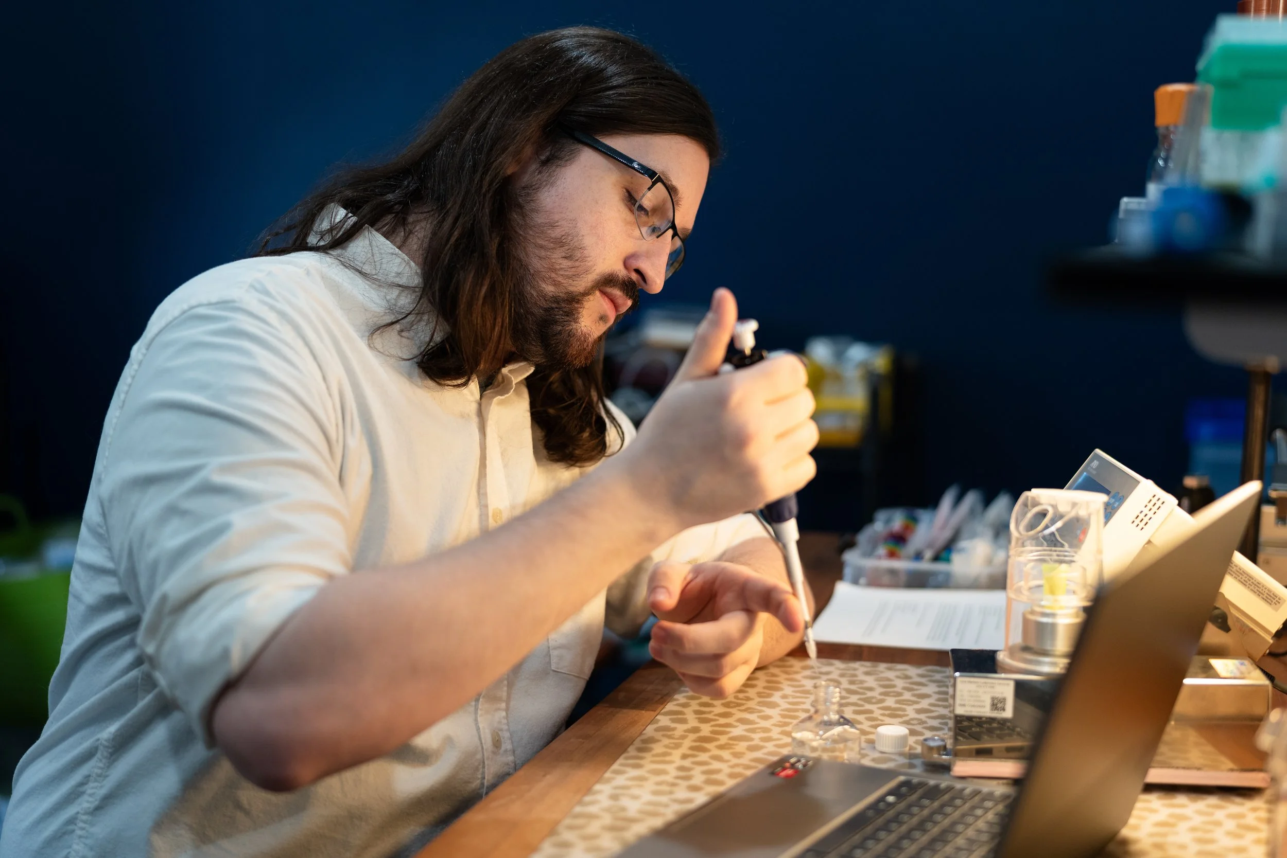 A man with long hair, beard, glasses, wearing a white shirt, working at a lab bench with scientific equipment, pipetting a liquid into a container, with a laptop and various lab tools around him.