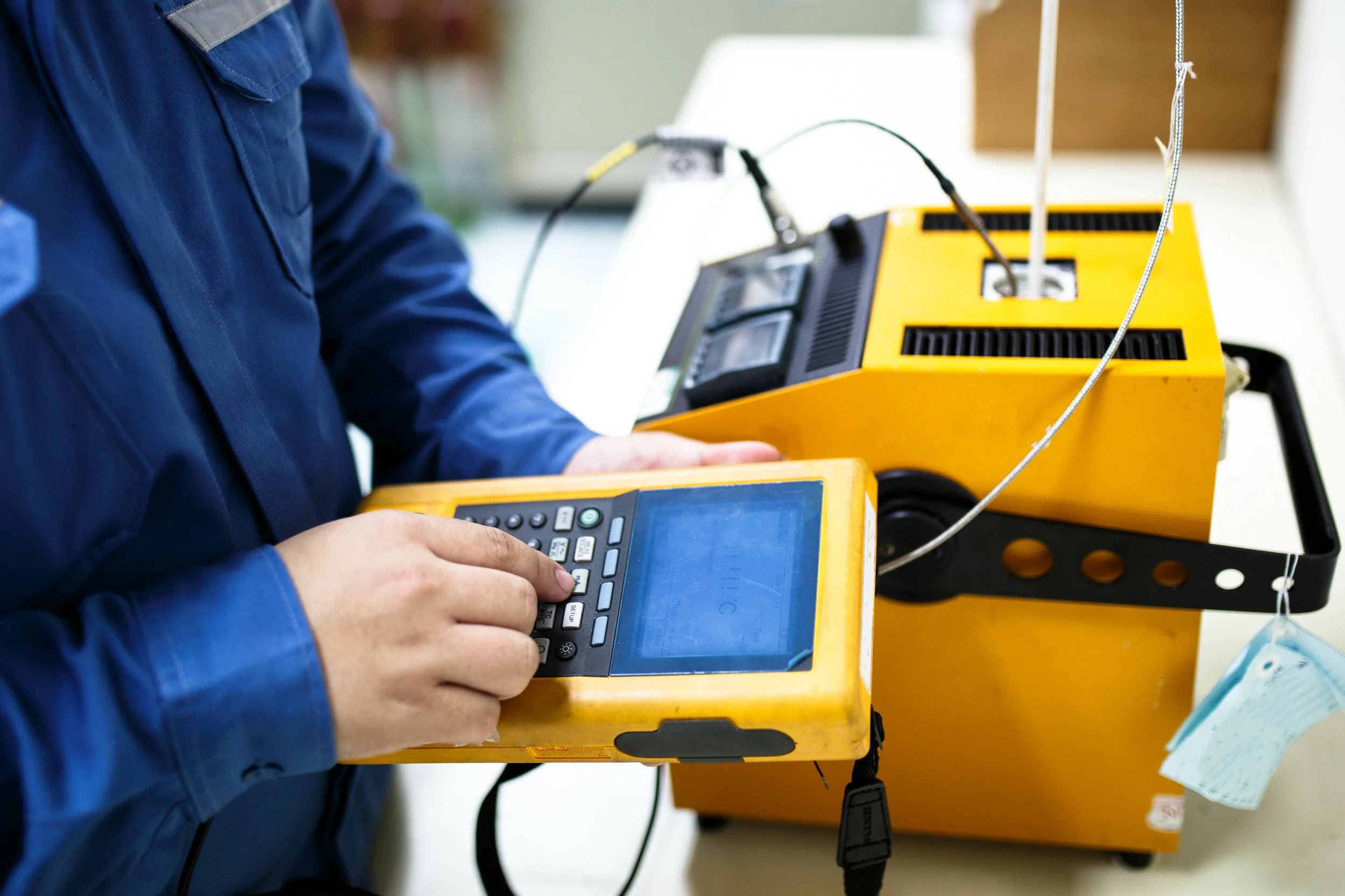 A person wearing a blue jacket is operating electronic testing equipment, with a yellow device and a multimeter or similar instrument on a white surface.
