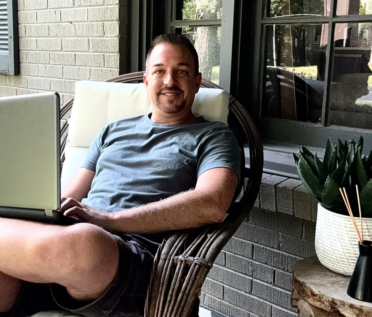 Man sitting on a wicker chair on a porch, smiling, with a laptop on his lap, next to a potted plant on a small side table.