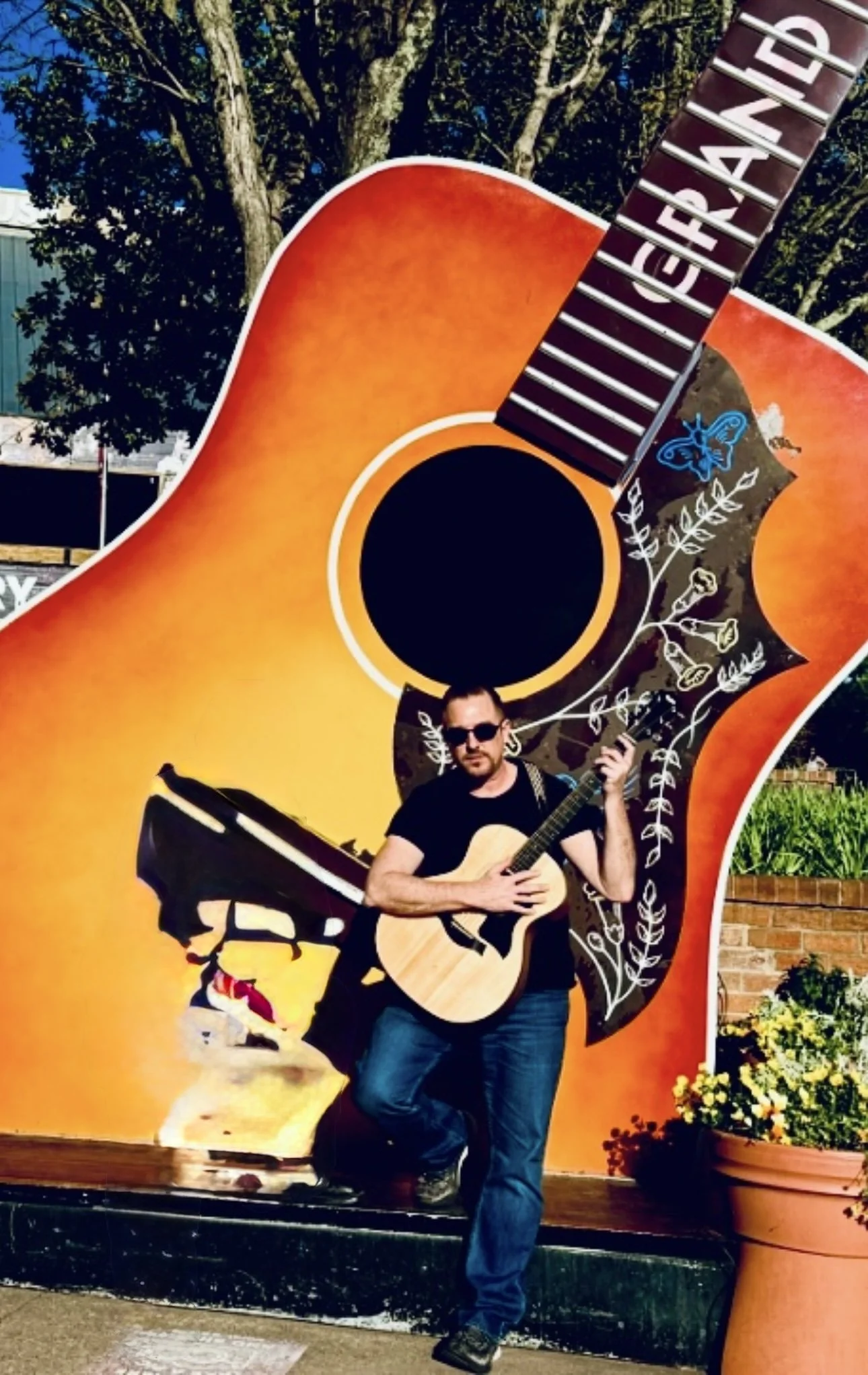 A man with sunglasses playing an acoustic guitar in front of a large colorful mural of a guitar.