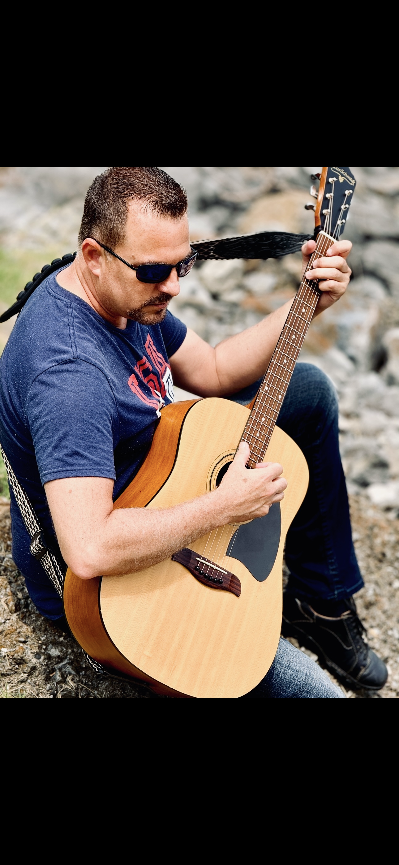 A man sitting on rocks outdoors playing an acoustic guitar, wearing sunglasses and a blue T-shirt.