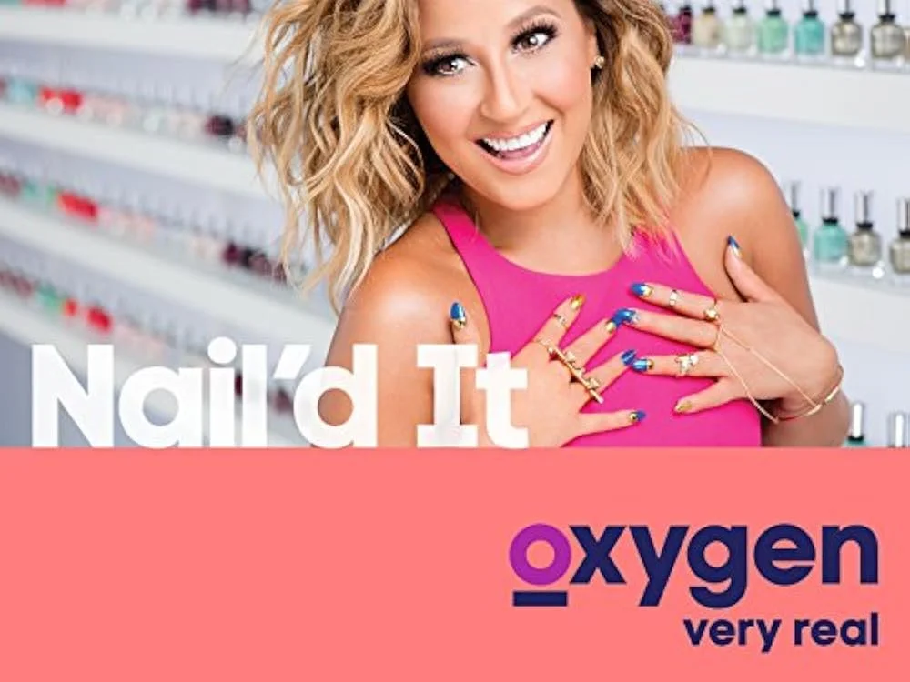 A woman with blonde curly hair smiling, showing her manicured nails, standing in front of a nail polish display. The text reads 'Nailed It' and 'oxygen very real'.