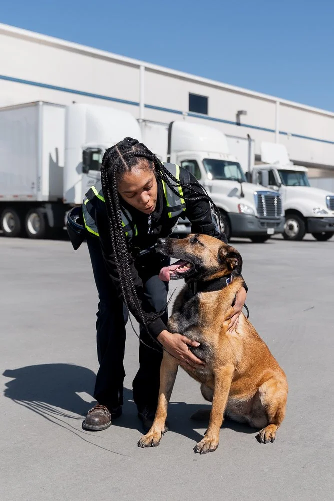 A GK9 handler kneels beside a Belgian Malinois detection canine, giving it an affectionate hold on a loading dock with trucks parked in the background under a bright blue sky.