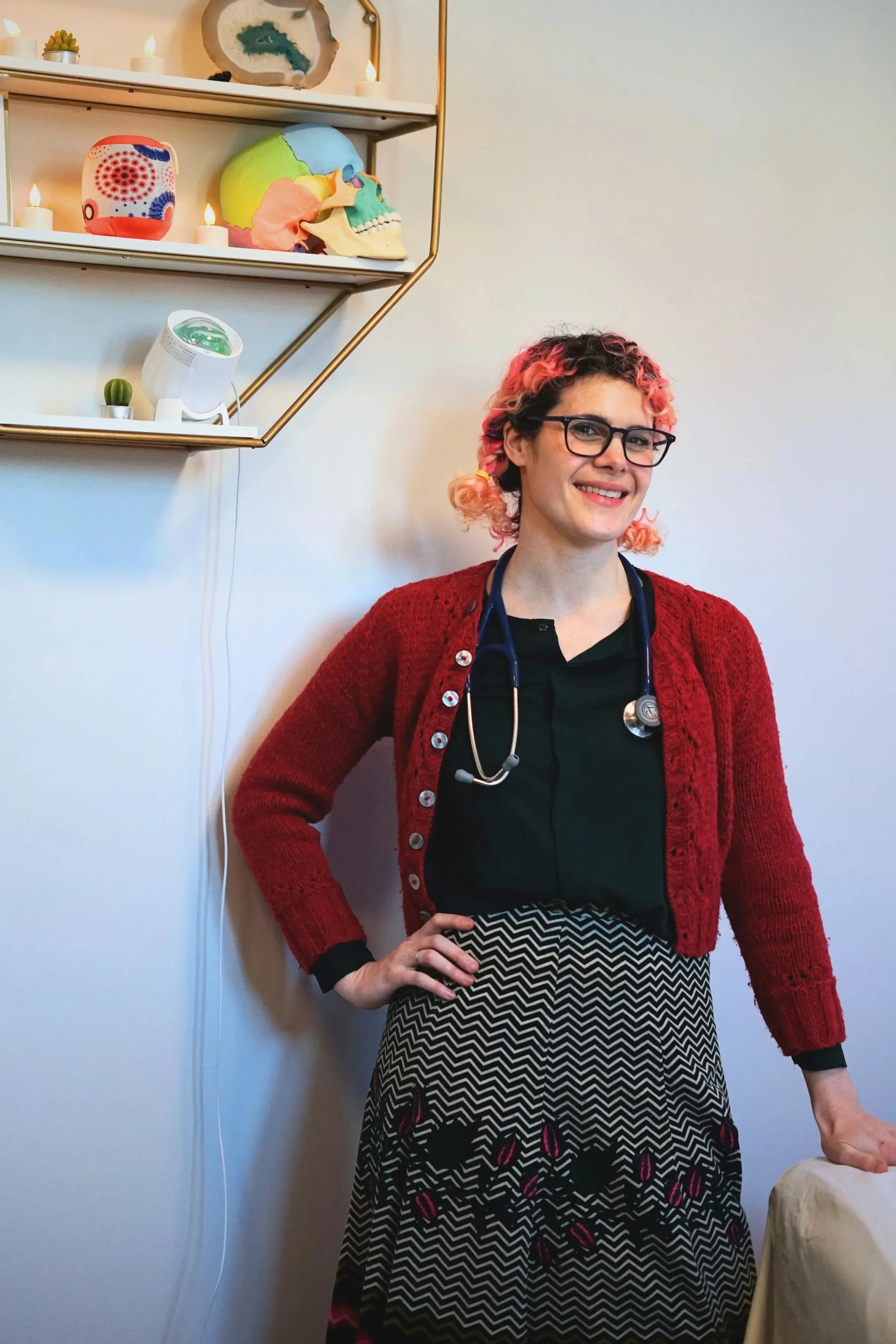 Dr Rodrigues with stethoscope, glasses and pink hair standing in her office in front of a shelf containing a model skull