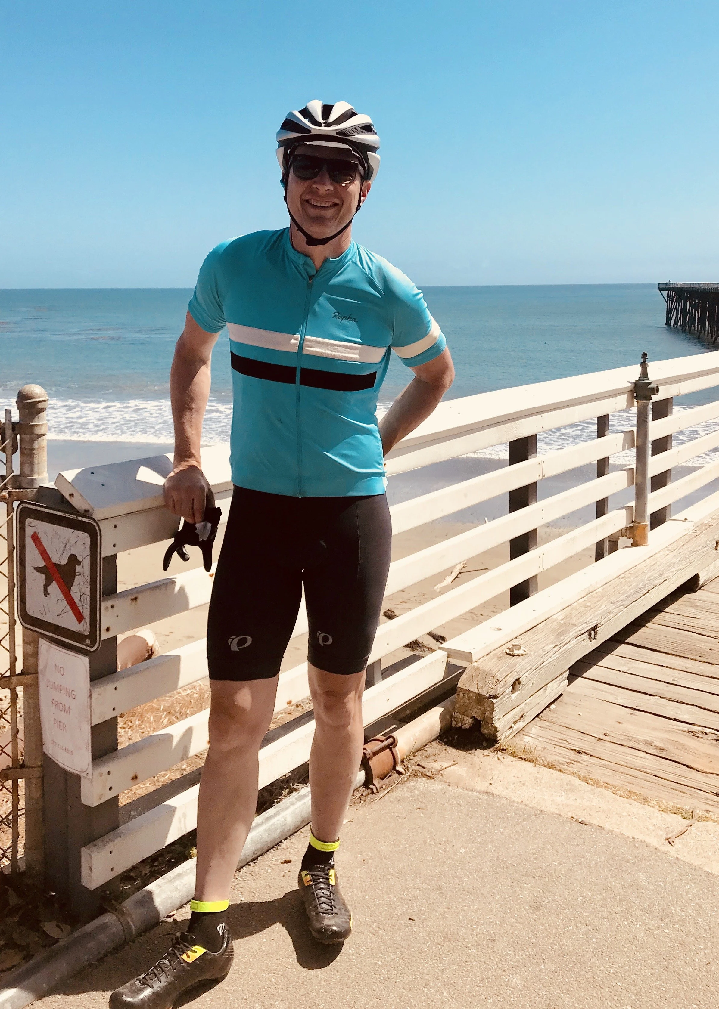 Man in cycling gear, wearing helmet and sunglasses, standing on a pier near the beach, smiling at camera.