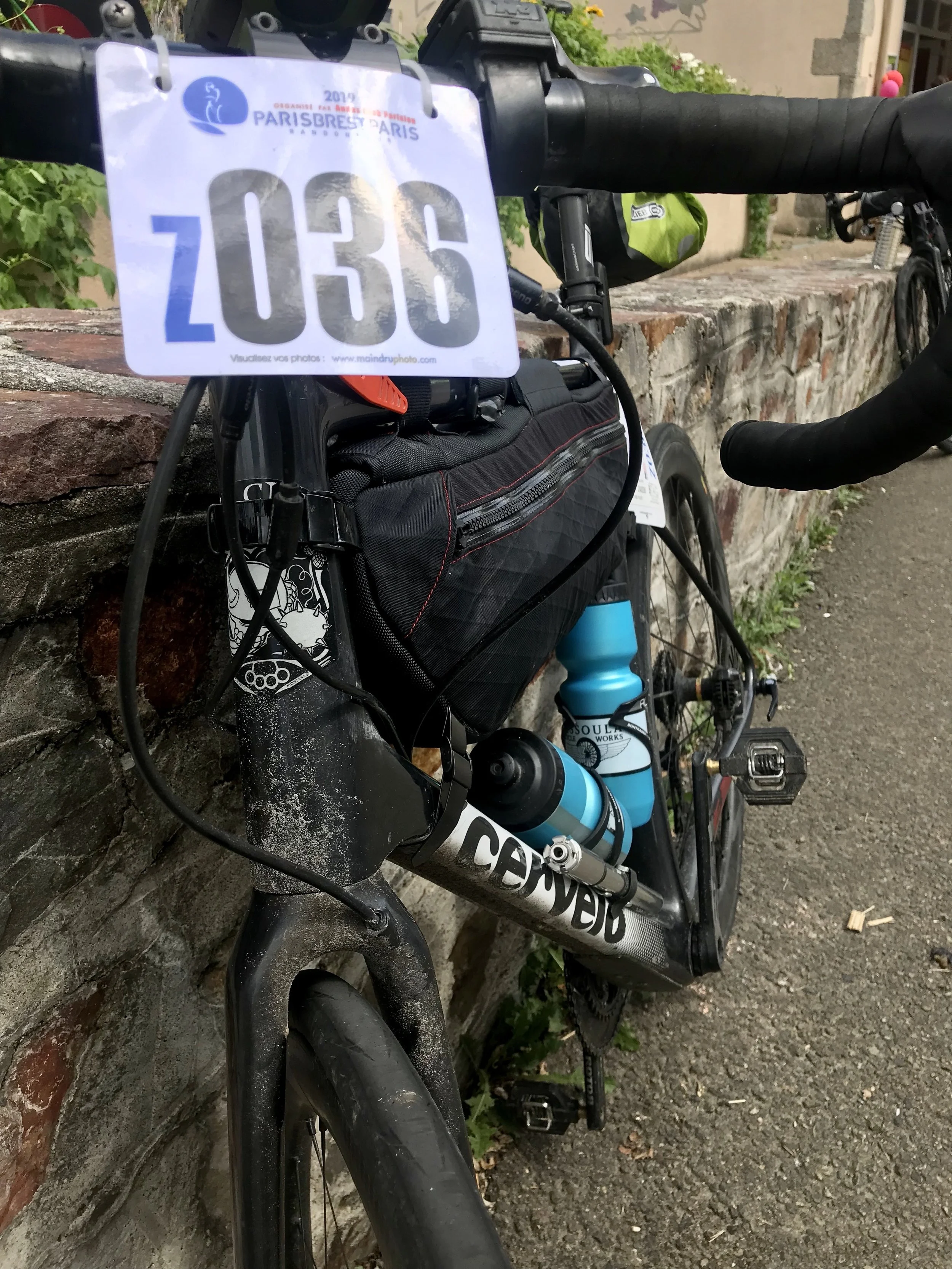 A black Trek bicycle with a blue water bottle, mounted on a brick wall during a cycling event, with a race number Z036 attached to the handlebars. Leif Bjelland Paris Brest Paris