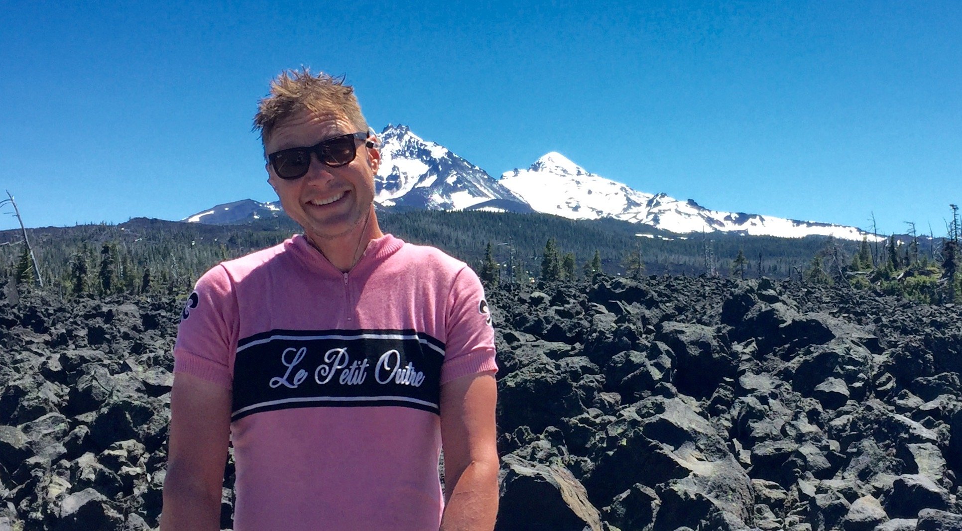A man wearing sunglasses and a pink cycling jersey smiling in front of a rocky landscape with snow-capped mountains and blue sky in the background. Leif Bjelland McKenzie Pass