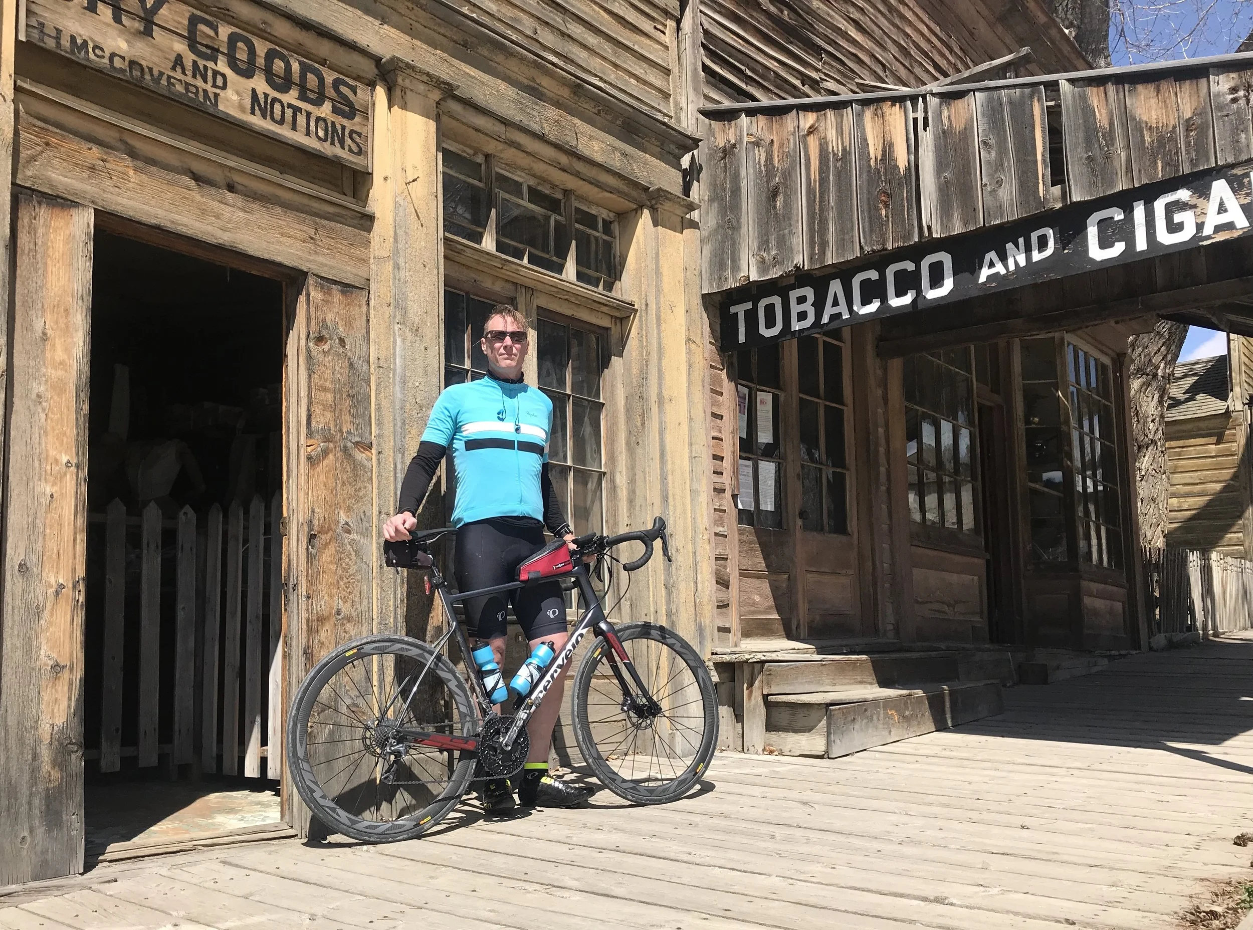 A man in cycling gear standing with a bicycle outside a wooden shop with signs that read "Tobacco and Cigar" and "Dry Goods and Grocery Notions".