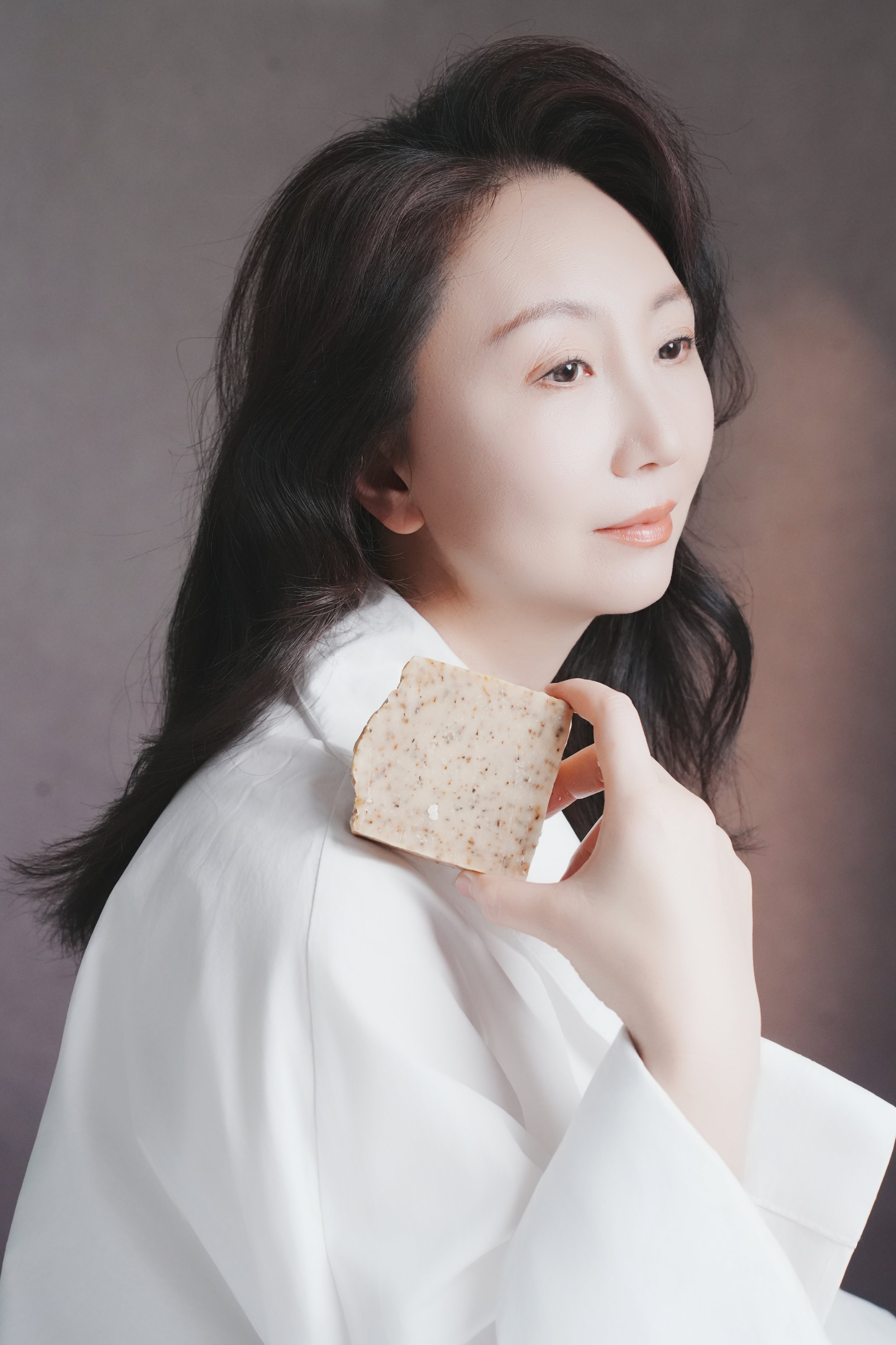 A woman with long dark hair wearing a white garment, holding a textured soap bar near her shoulder, with a neutral background.