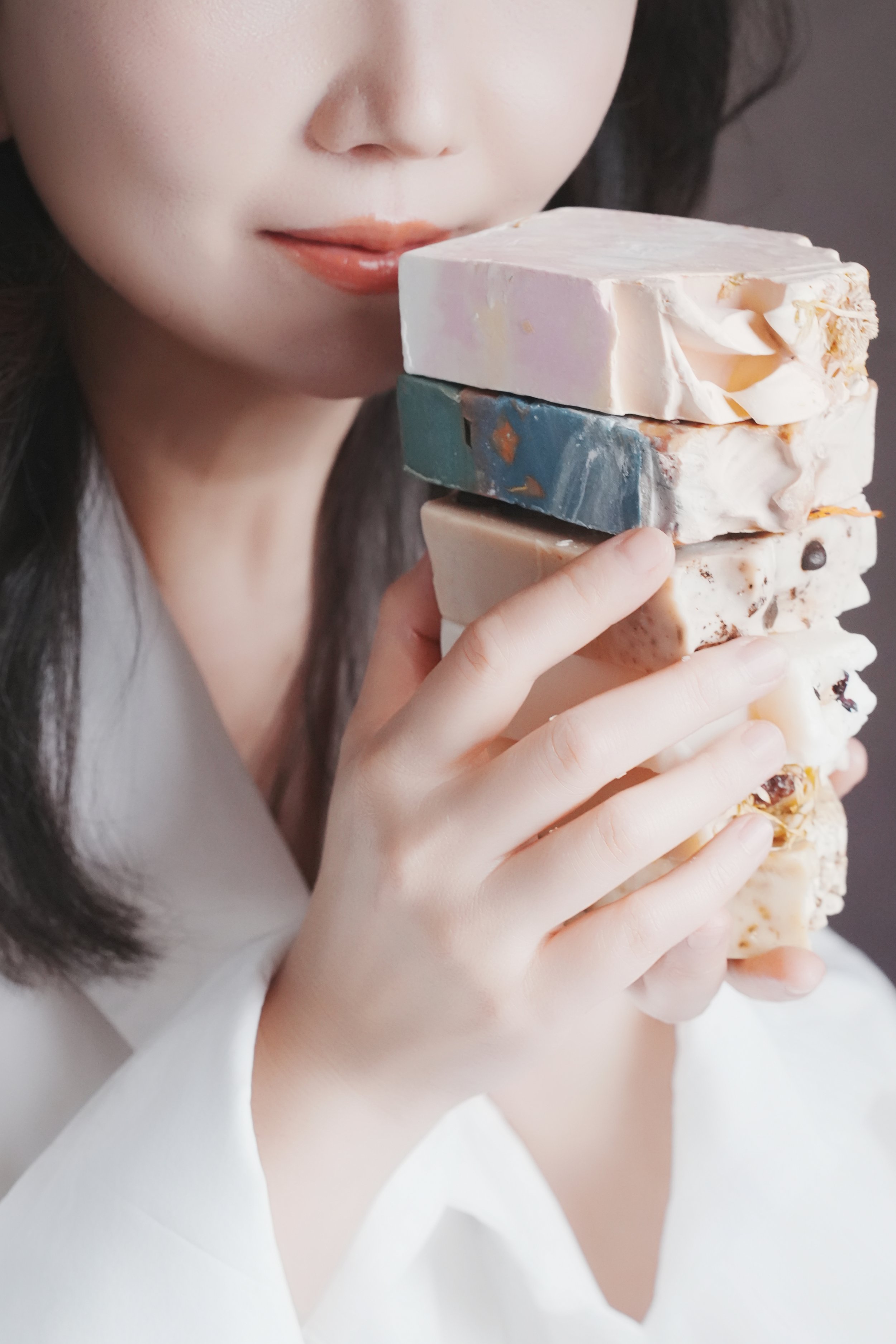 A woman with dark hair and fair skin holding a stack of various artisanal soap bars.