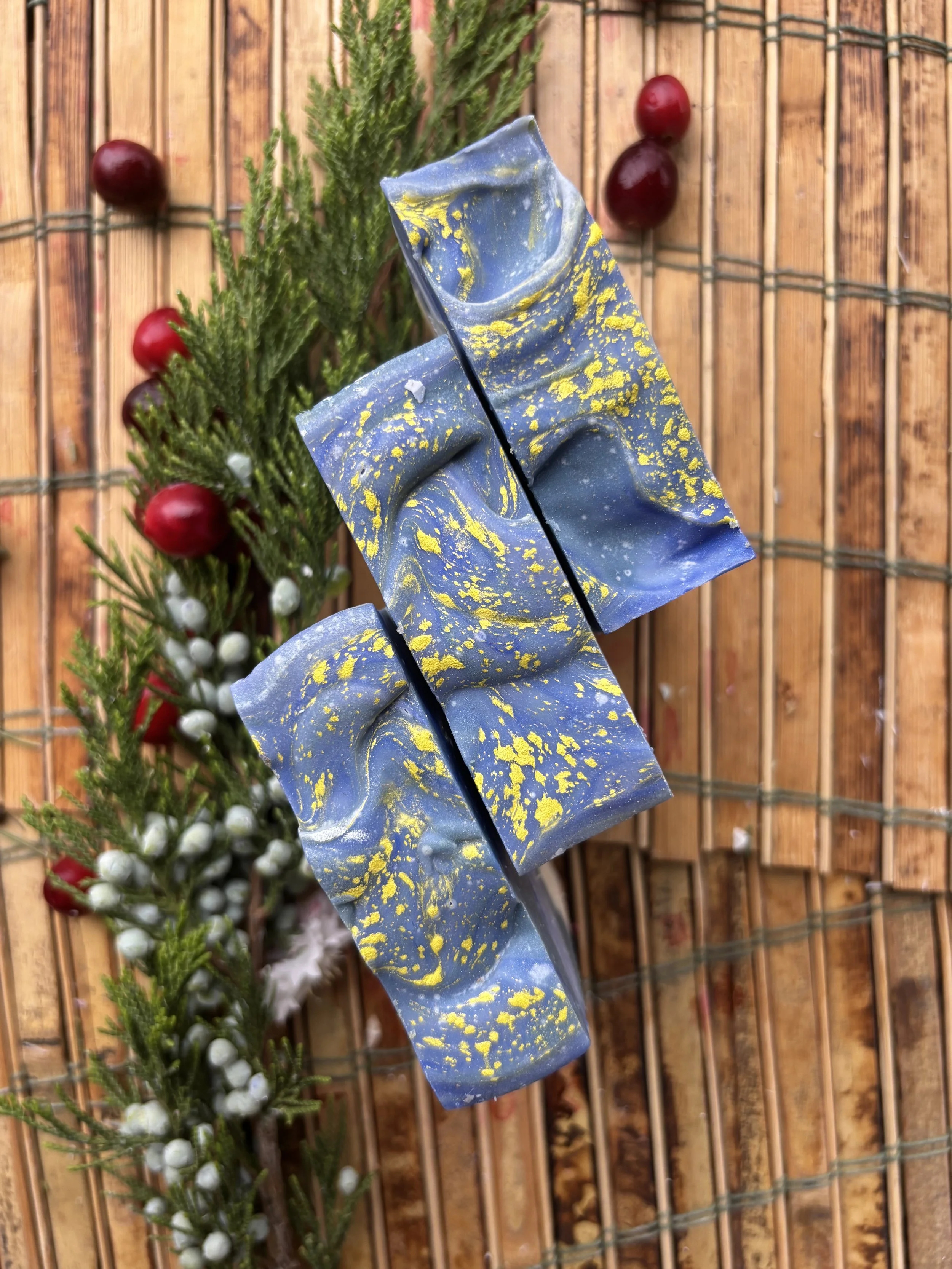 Three artistically painted soap bars with blue and yellow paint on a wooden surface, alongside greenery and red berries.