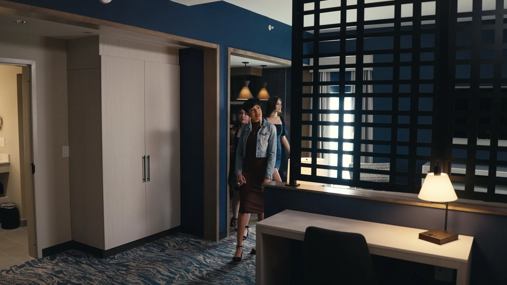 Three women walking into a hotel room, with dark blue walls, a white closet door, and a desk lamp in the foreground.