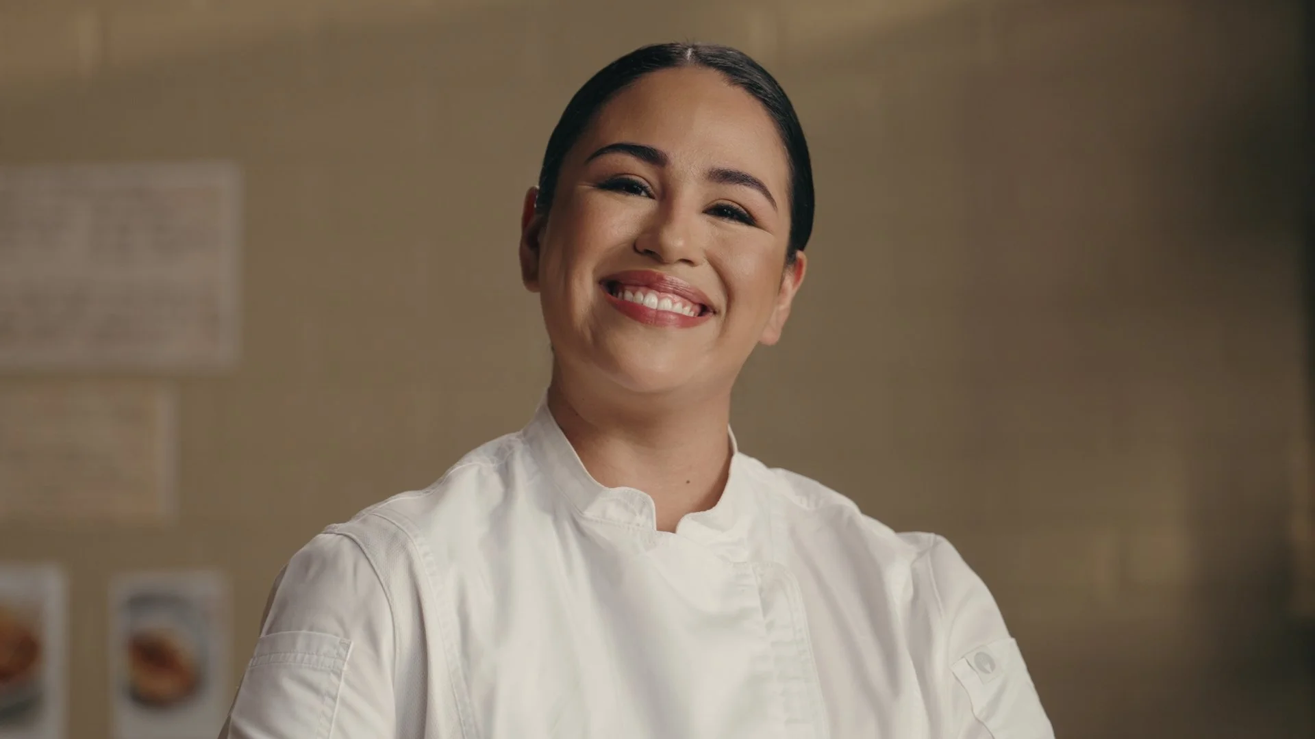 Smiling woman chef with dark hair, wearing a white chef jacket, standing in a kitchen