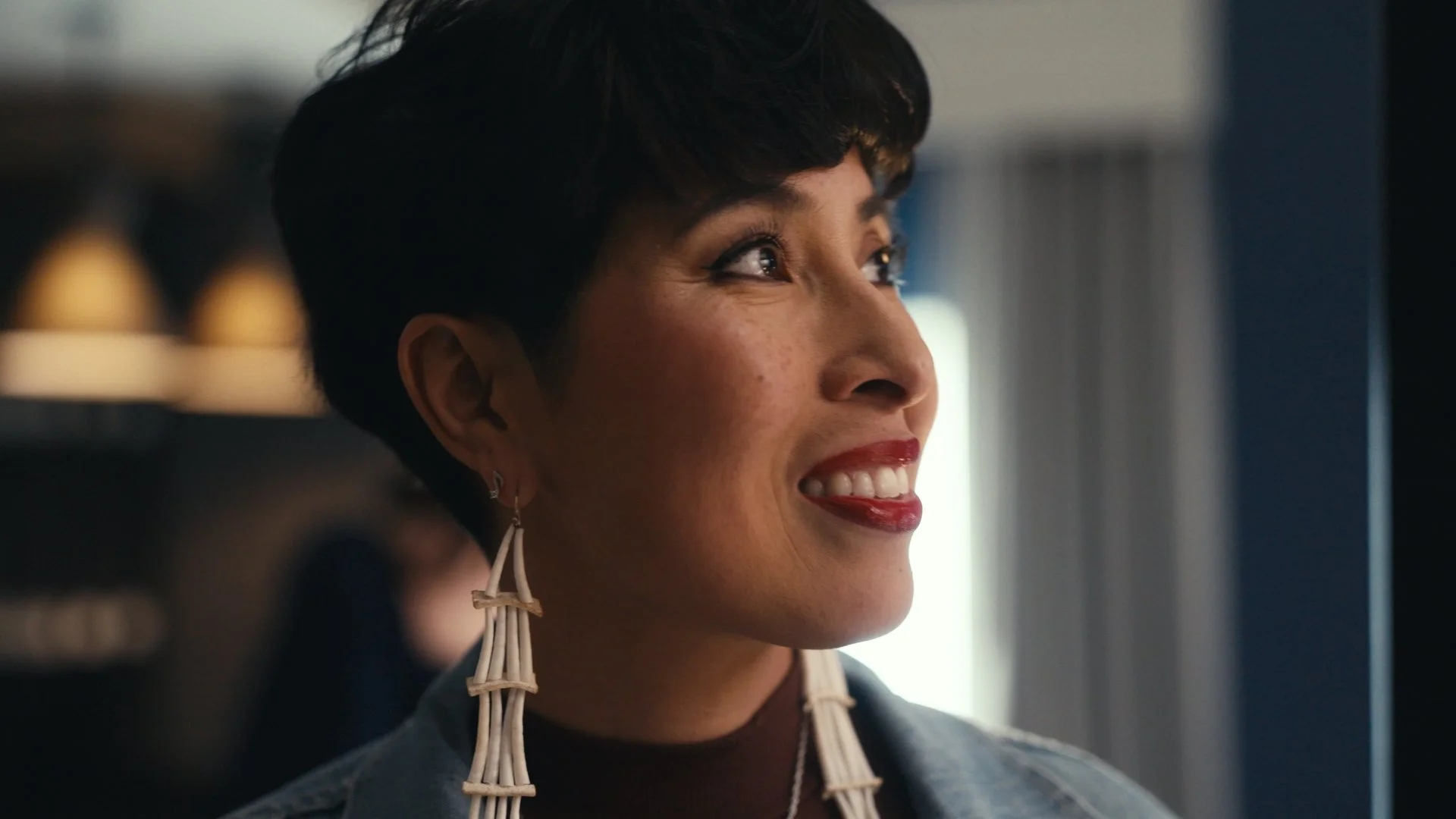 A woman with short dark hair, wearing statement earrings and a denim jacket, smiling and looking upwards indoors.