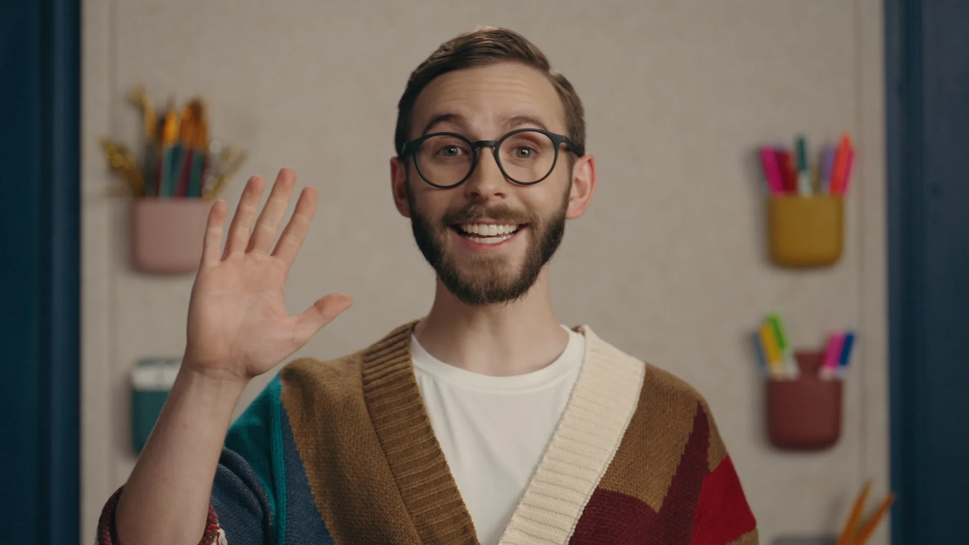 Smiling man with glasses waving indoors, background with colorful wall organizers holding pens and markers.