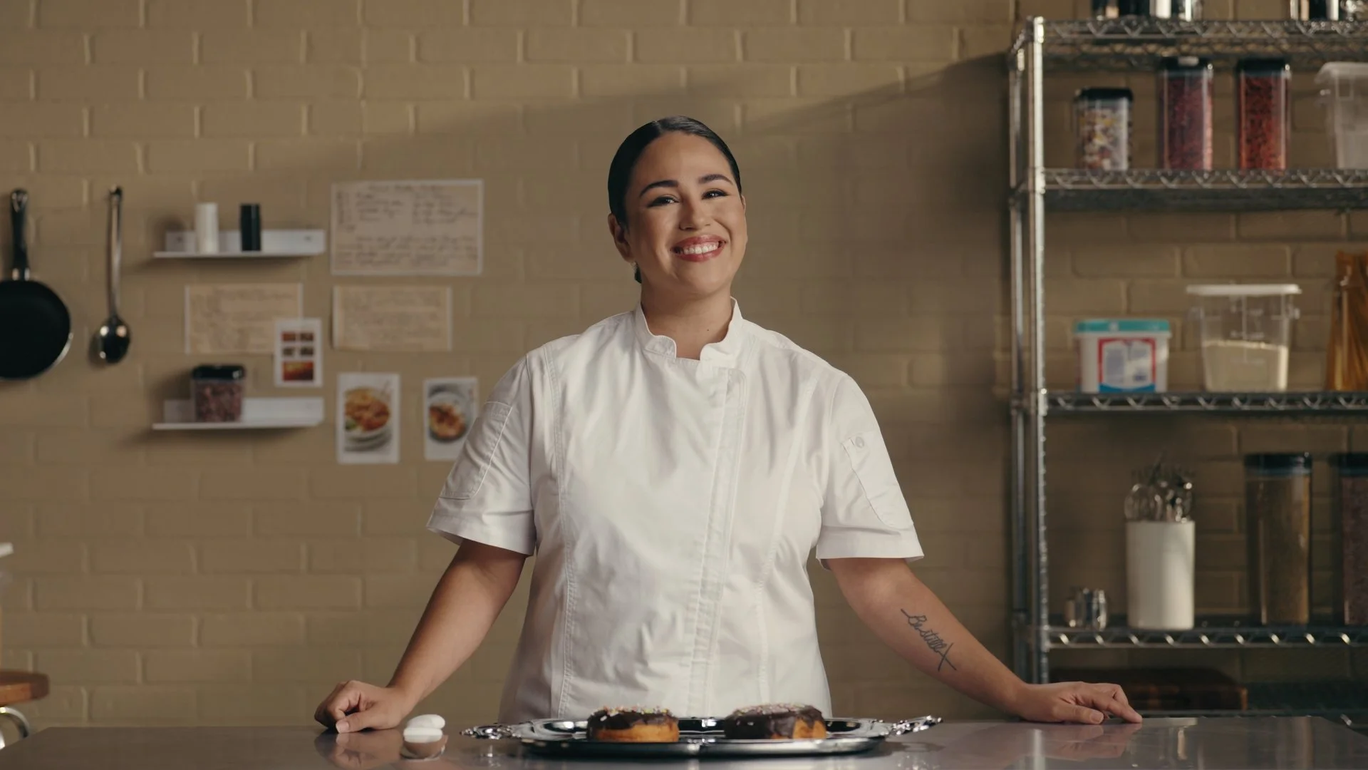A smiling female chef standing behind a kitchen counter with two éclairs on a silver platter.