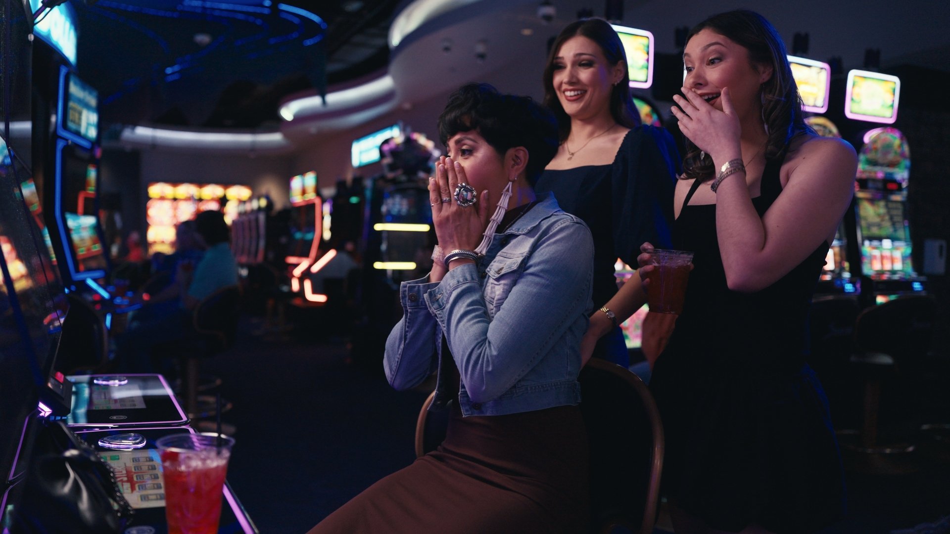 Three women at a casino looking excited and surprised while playing slot machines, with colorful machines in the background.