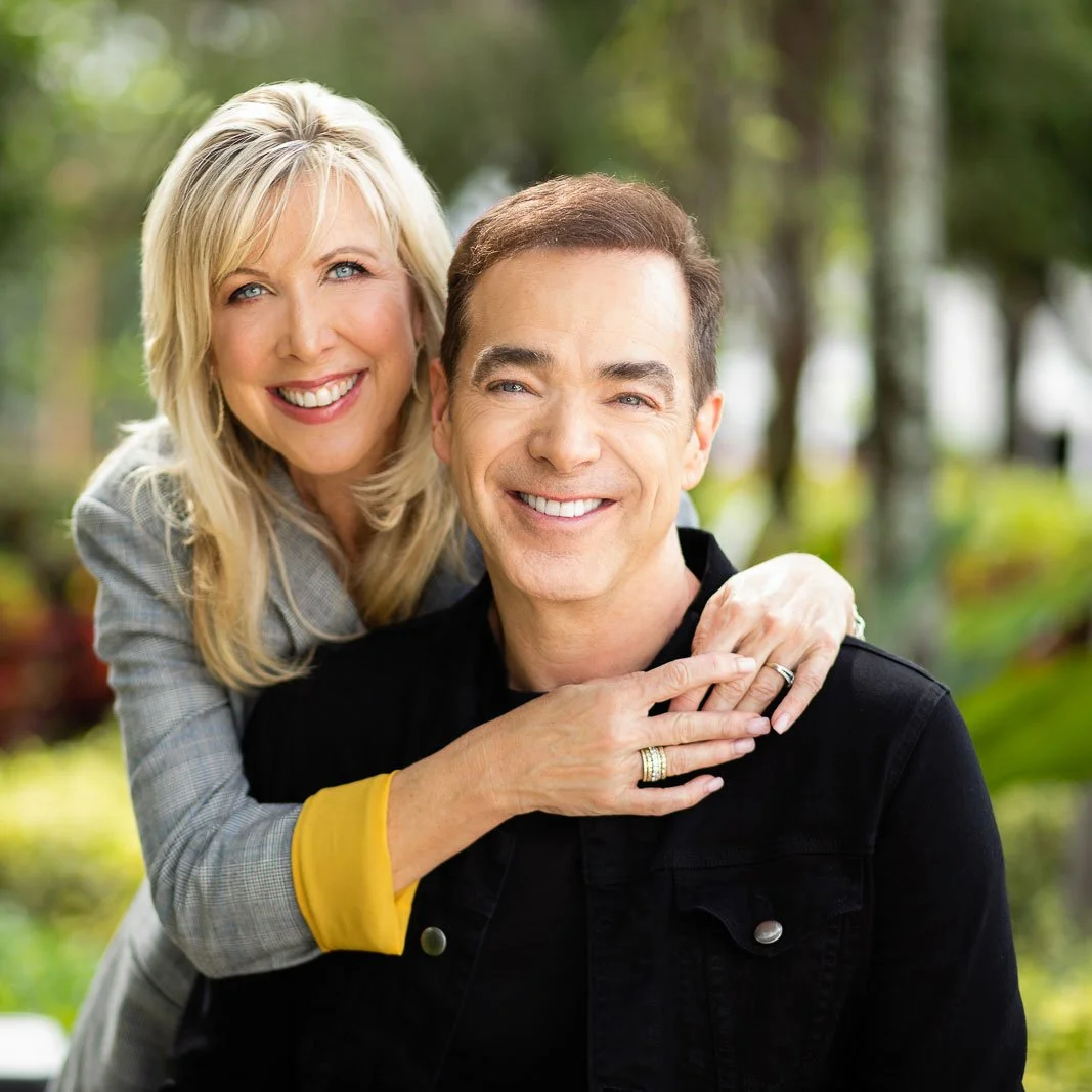 A smiling woman with blonde hair embraces a man with dark hair from behind in a park with green trees.