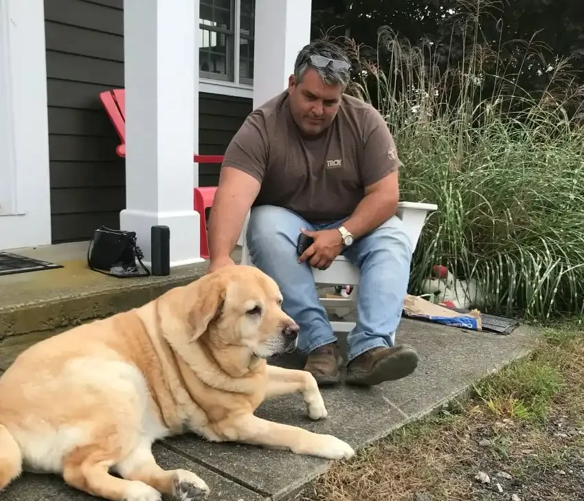 A man sitting on a porch next to a large Labrador Retriever dog.