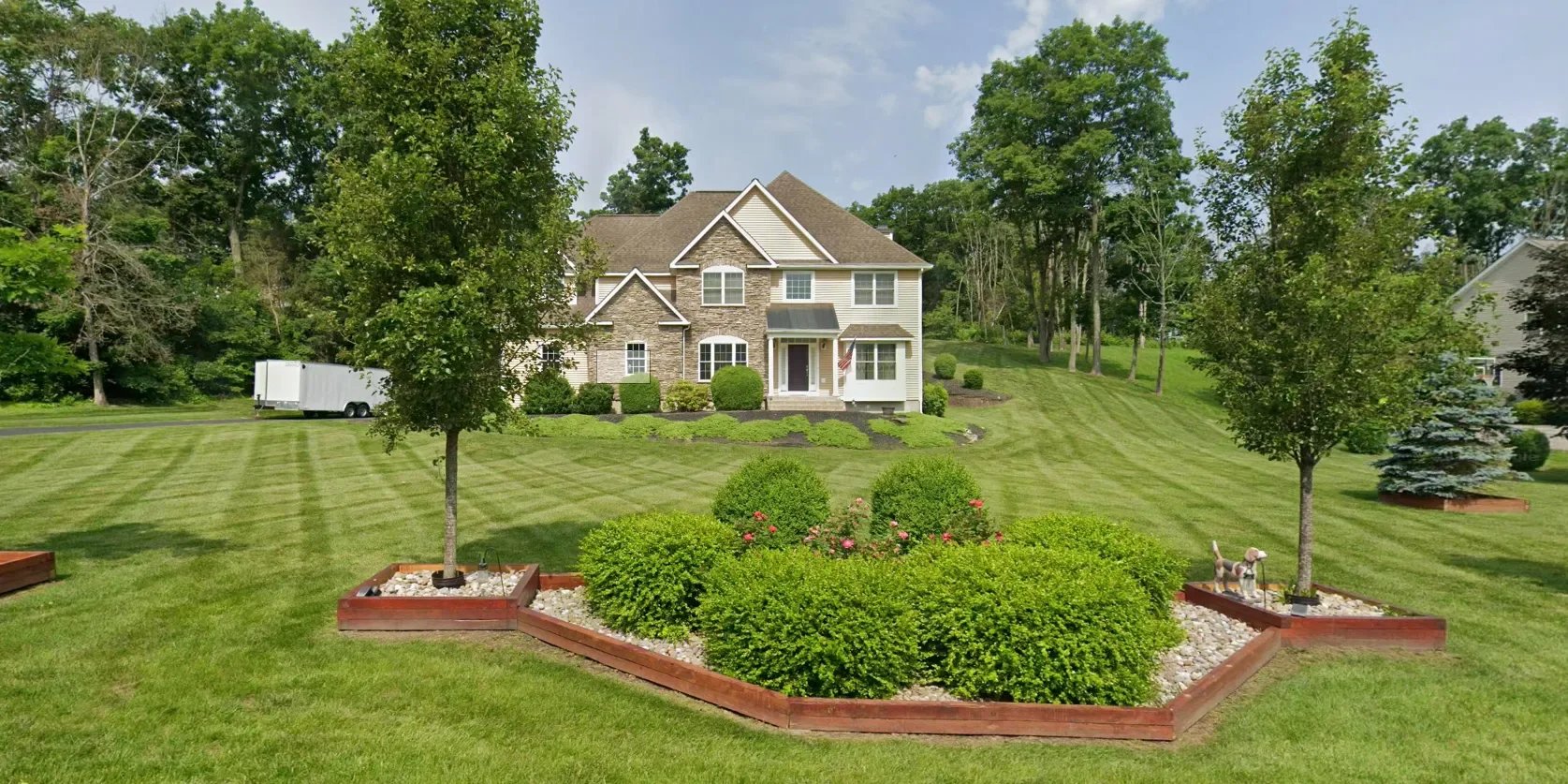 Beautiful front yard with well-maintained grass, two small trees in decorated flower beds, and a large house with stone and siding exterior in the background, surrounded by green trees.