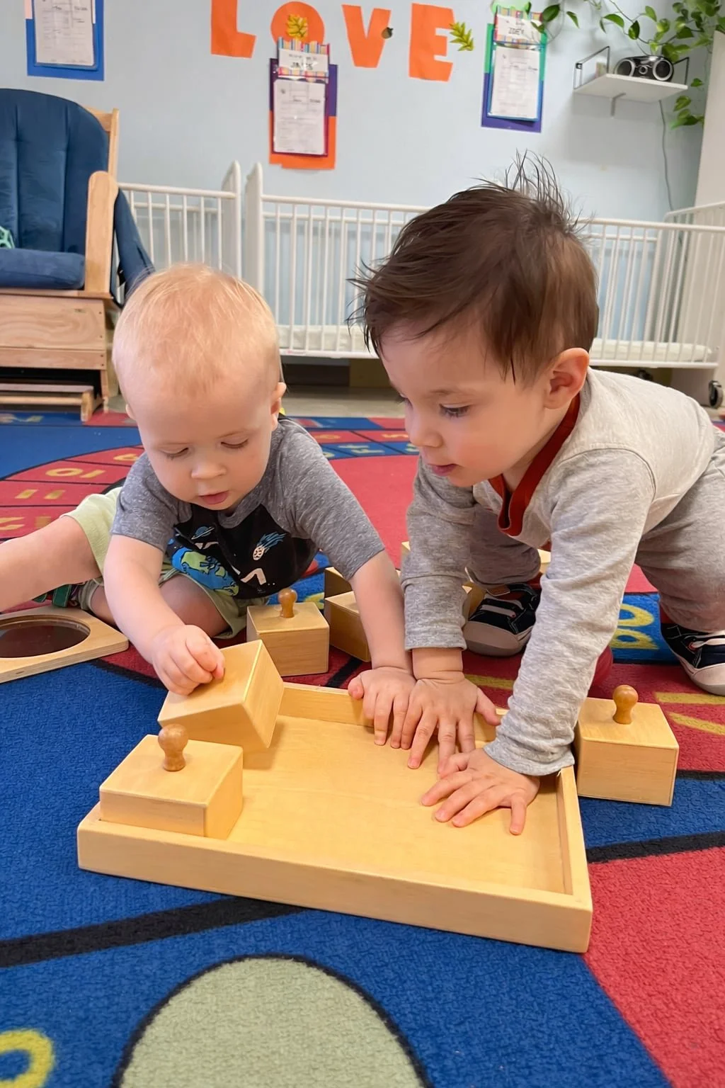 Two infants enjoy Montessori wooden puzzles on a rug in an Austin Montessori preschool in Texas.