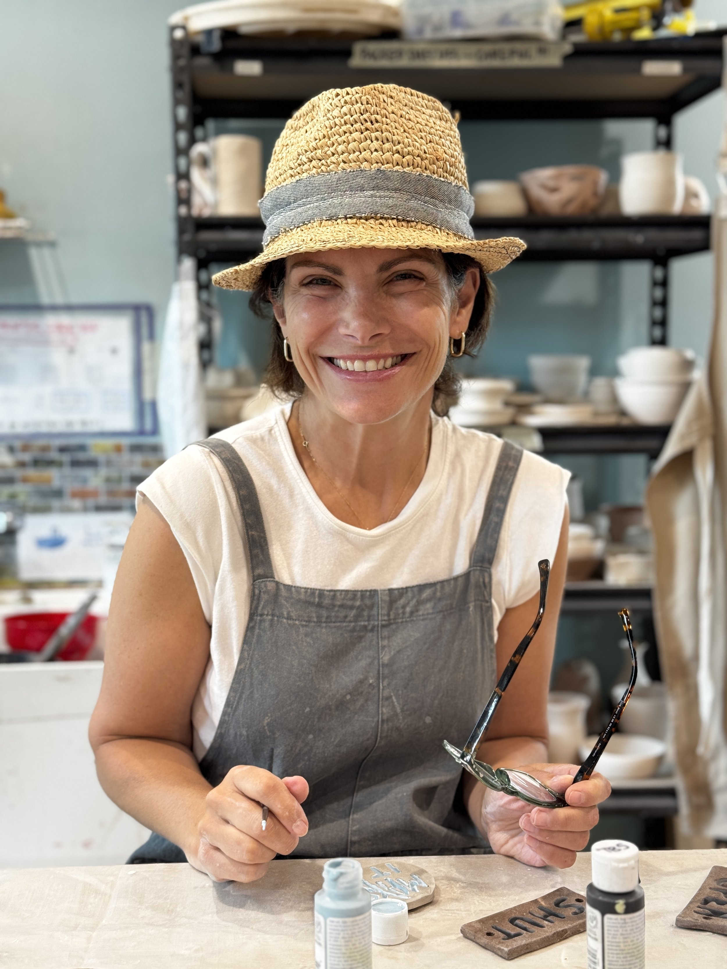 Woman in a straw hat and gray apron holding glasses, working on a craft or art project in a studio with shelves of pottery and art supplies in the background.