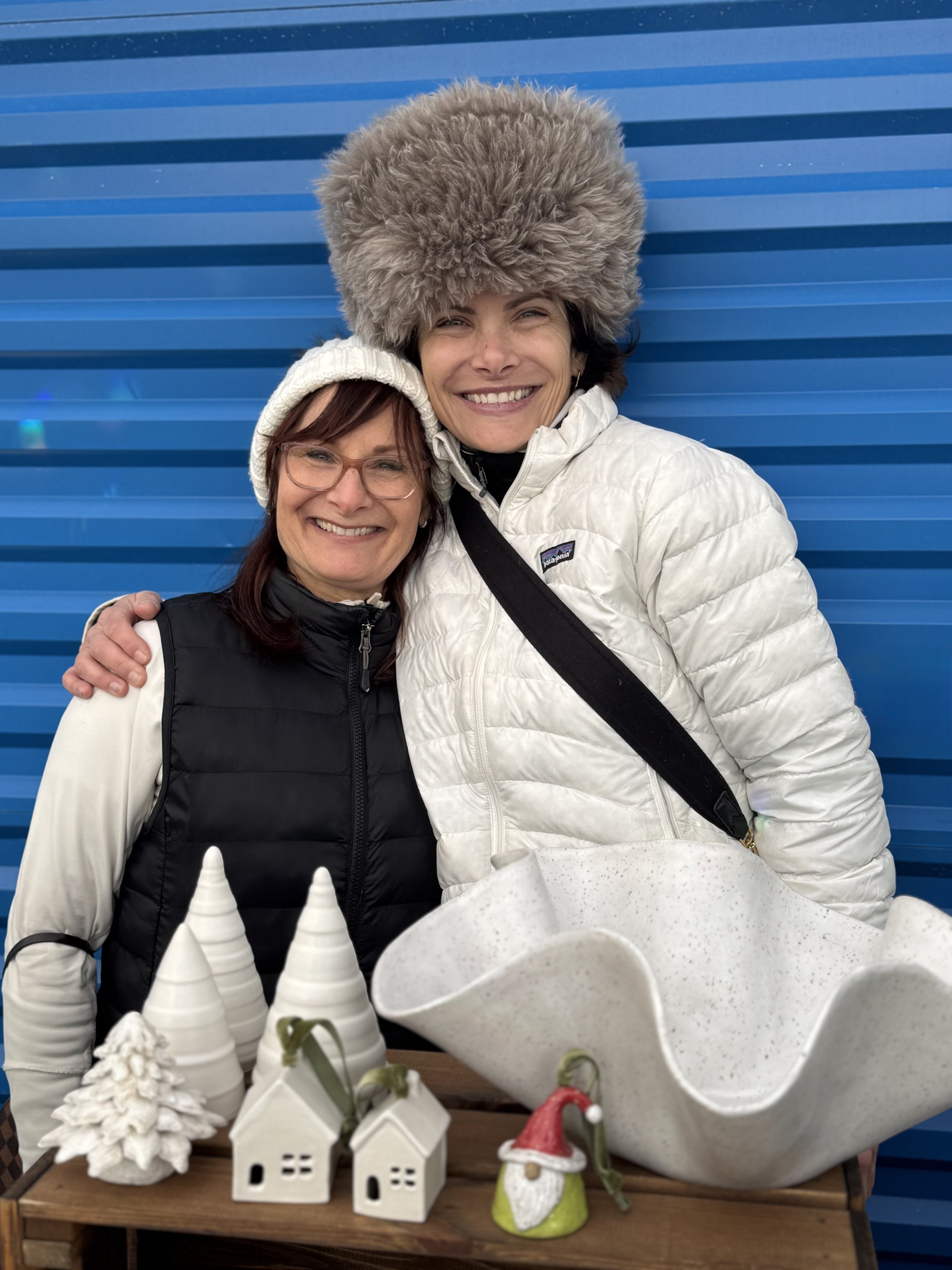 Two women smiling for a photo, standing behind a table with ceramic Christmas decorations including snow-covered trees, houses, a gnome, and a Christmas hat, in front of a blue ribbed wall.