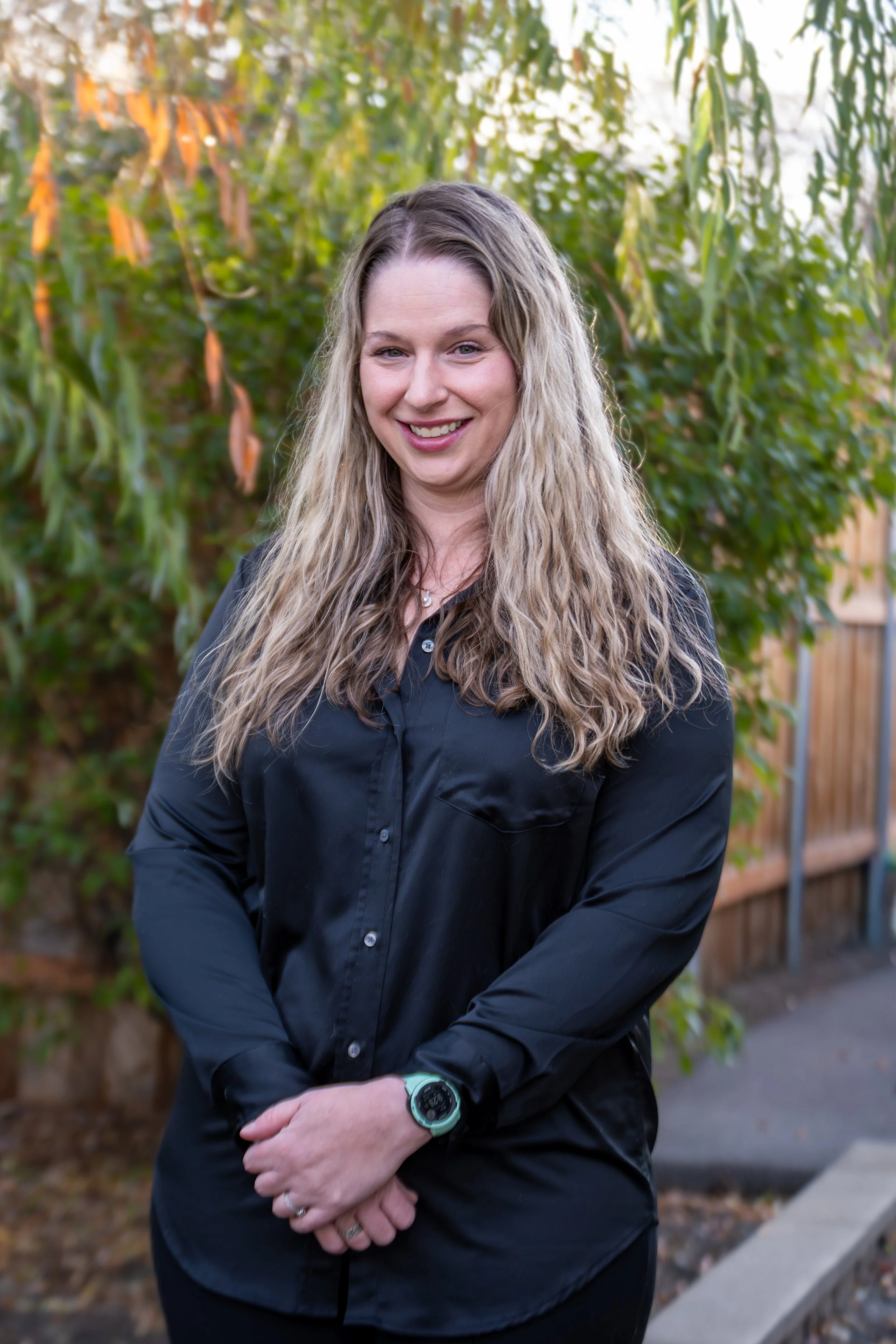 A woman standing outdoors in front of greenery, smiling, wearing a dark shirt and a watch.