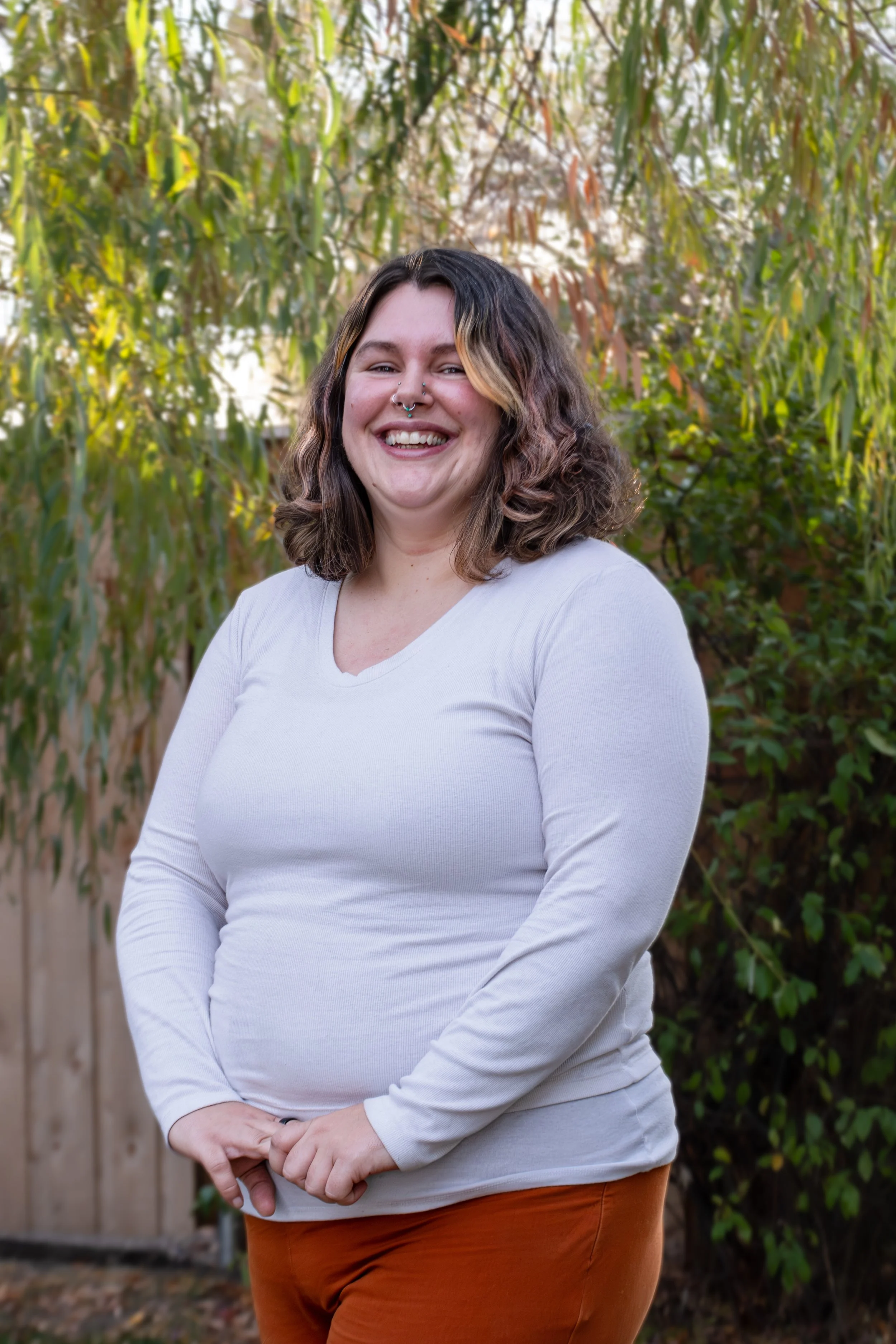 A woman smiling outdoors with trees and greenery in the background, wearing a white long-sleeve shirt and brown pants.