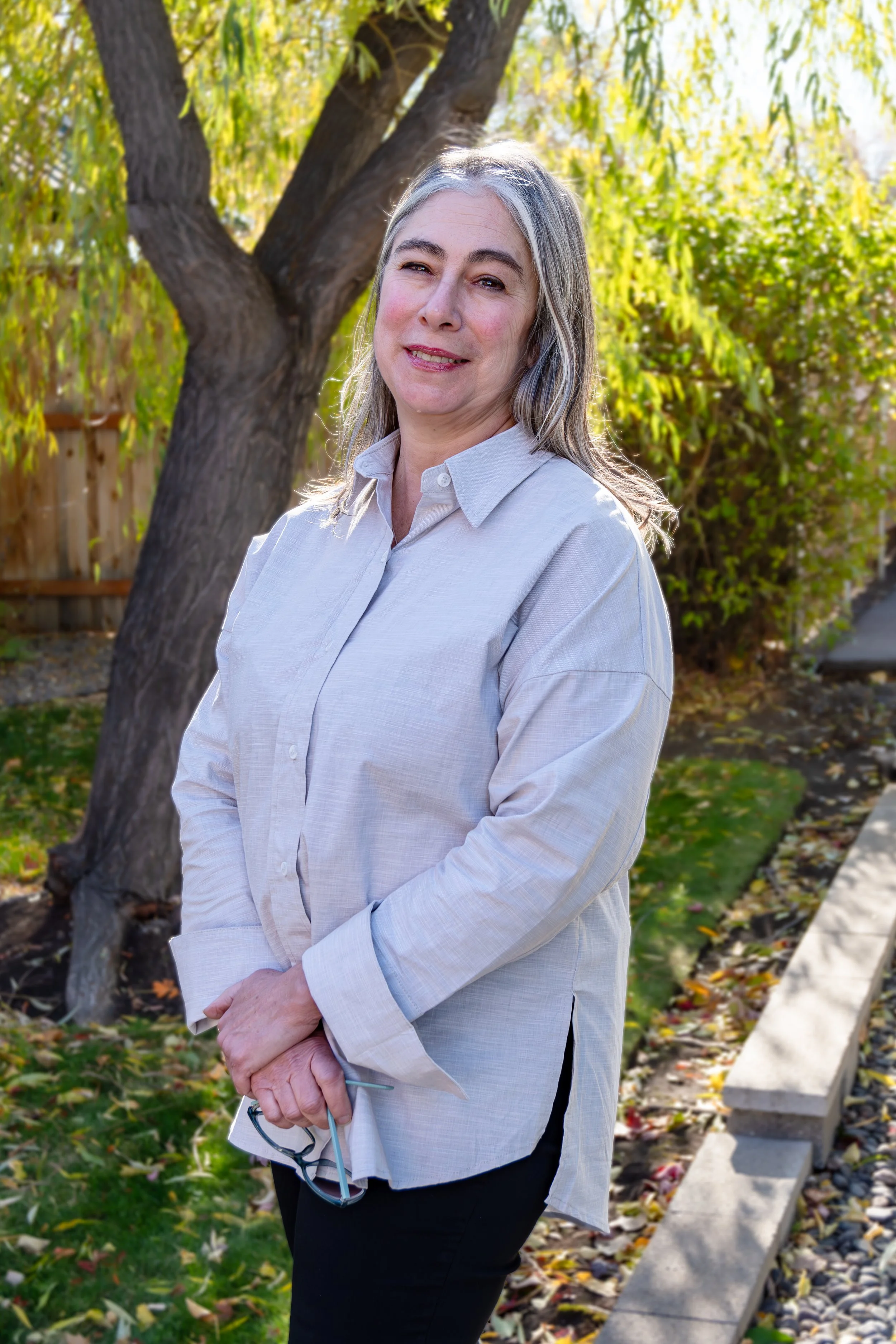 An older woman with long gray hair, wearing a white button-up shirt, standing outdoors near a tree with green leaves, in a landscaped garden setting.