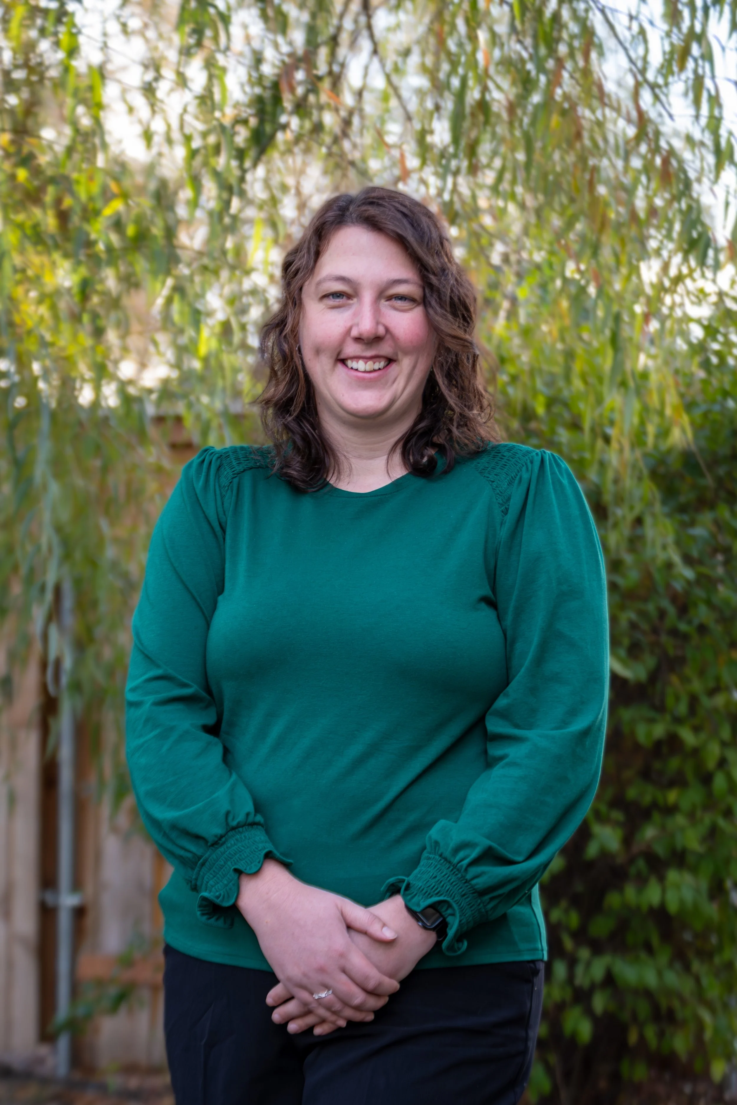 A woman with shoulder-length curly hair smiling outdoors, wearing a teal long-sleeve top with greenery in the background.