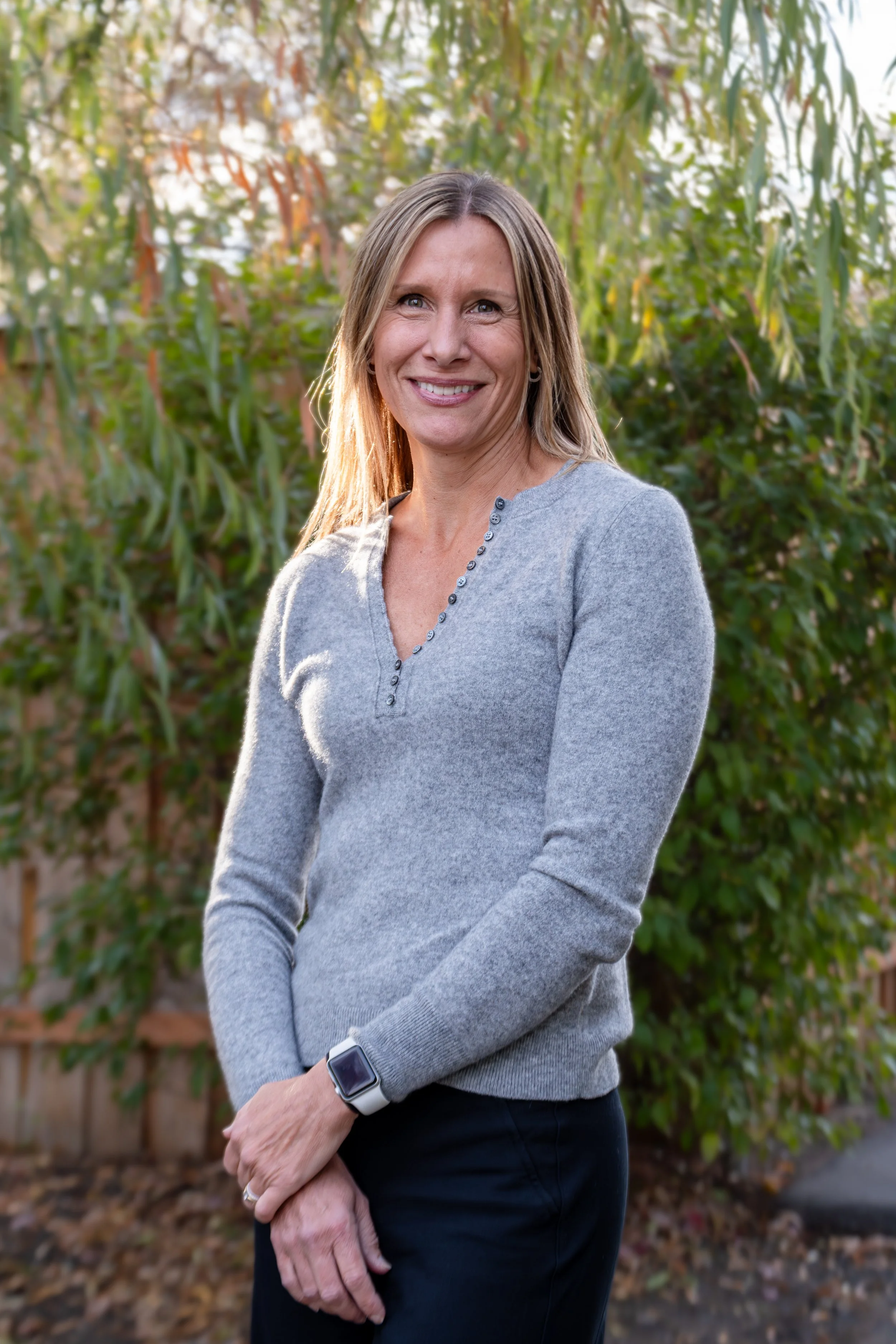 A woman standing outdoors in a natural setting with trees and foliage in the background, smiling at the camera.