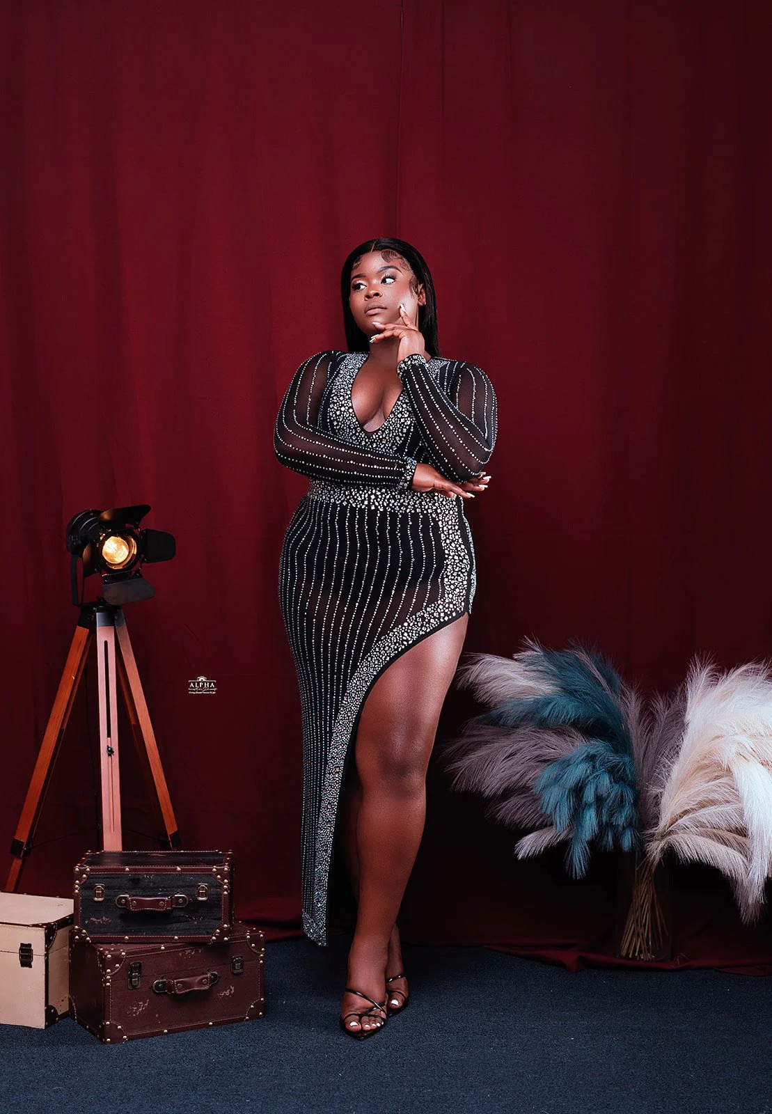 A woman in a black, bejeweled dress with a high slit poses with her hand on her chin against a red backdrop, with vintage-style suitcases, a tripod, and fluffy decorative plants nearby.